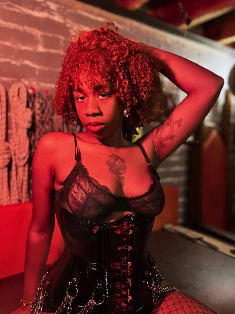 A woman with curly red hair wearing a black lace top and a shiny black corset with chains, sitting with one arm raised behind her head, in a dimly lit room with red lighting.