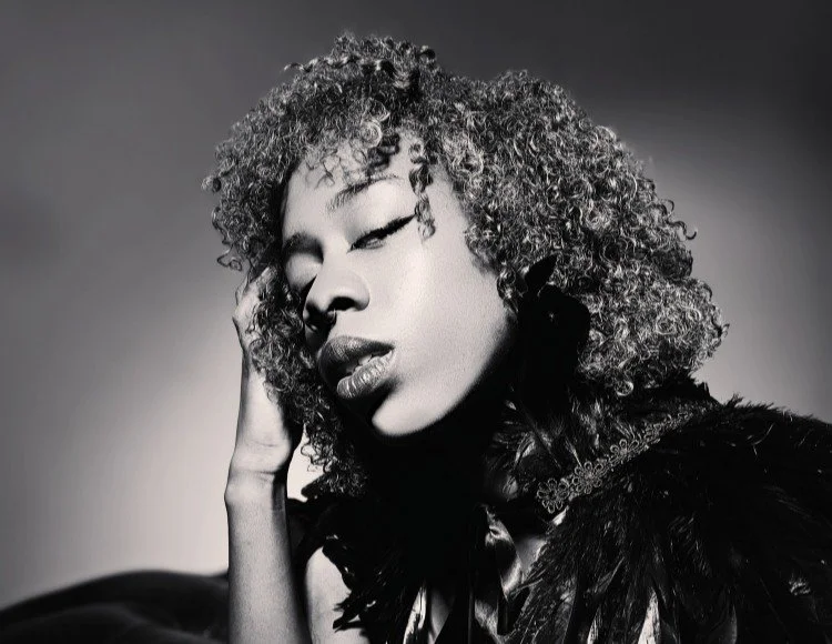 A black and white portrait of a woman with curly hair, closed eyes, and lips slightly parted, touching her face with her hand, wearing a dark, feathered or textured garment.