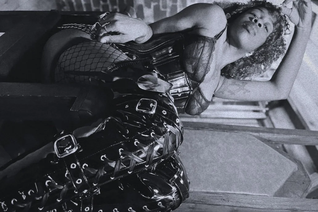 A woman with curly hair lying on a bed or couch, wearing a black lace top and fishnet stockings, with a black leather bag or purse nearby.