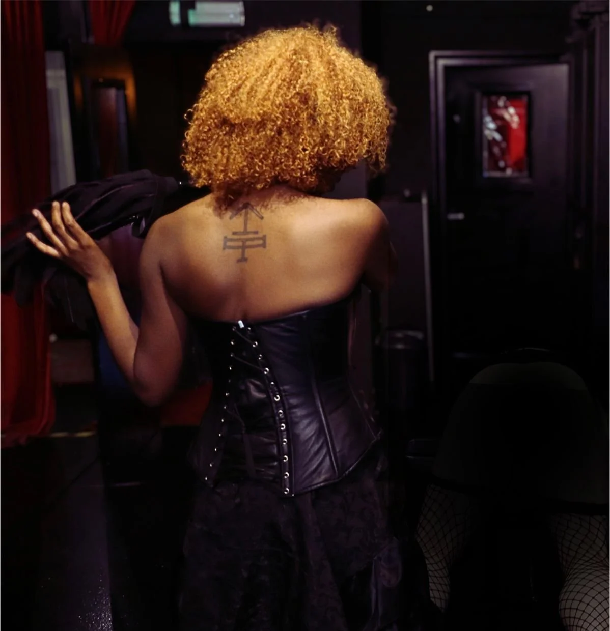 A woman with curly blonde hair wearing a black leather corset, showing a chest tattoo, in a dark room.