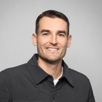 A man with short dark hair, wearing a black collared shirt and smiling, appears against a light gray background.
