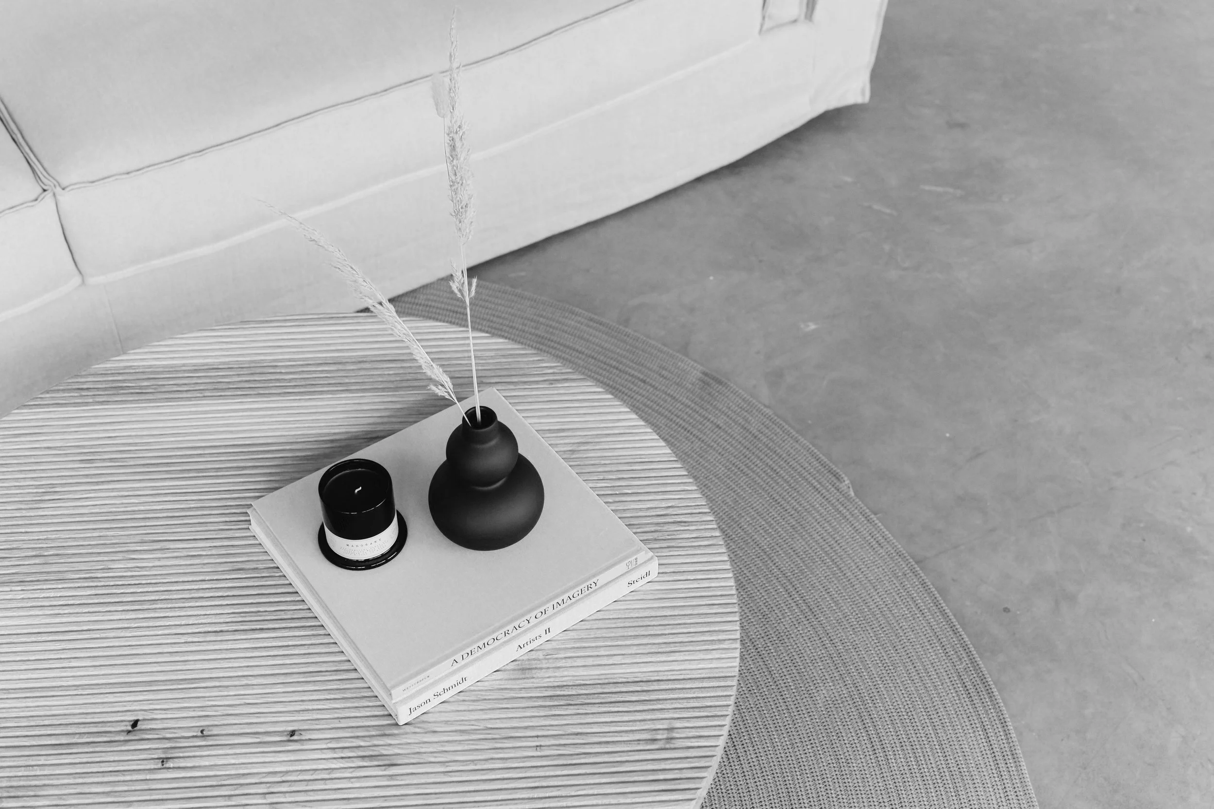 Top view of a round wooden table with a black vase holding dried grass, a black candle, and a book titled 'A Democracy of Imagery' in a modern living room.