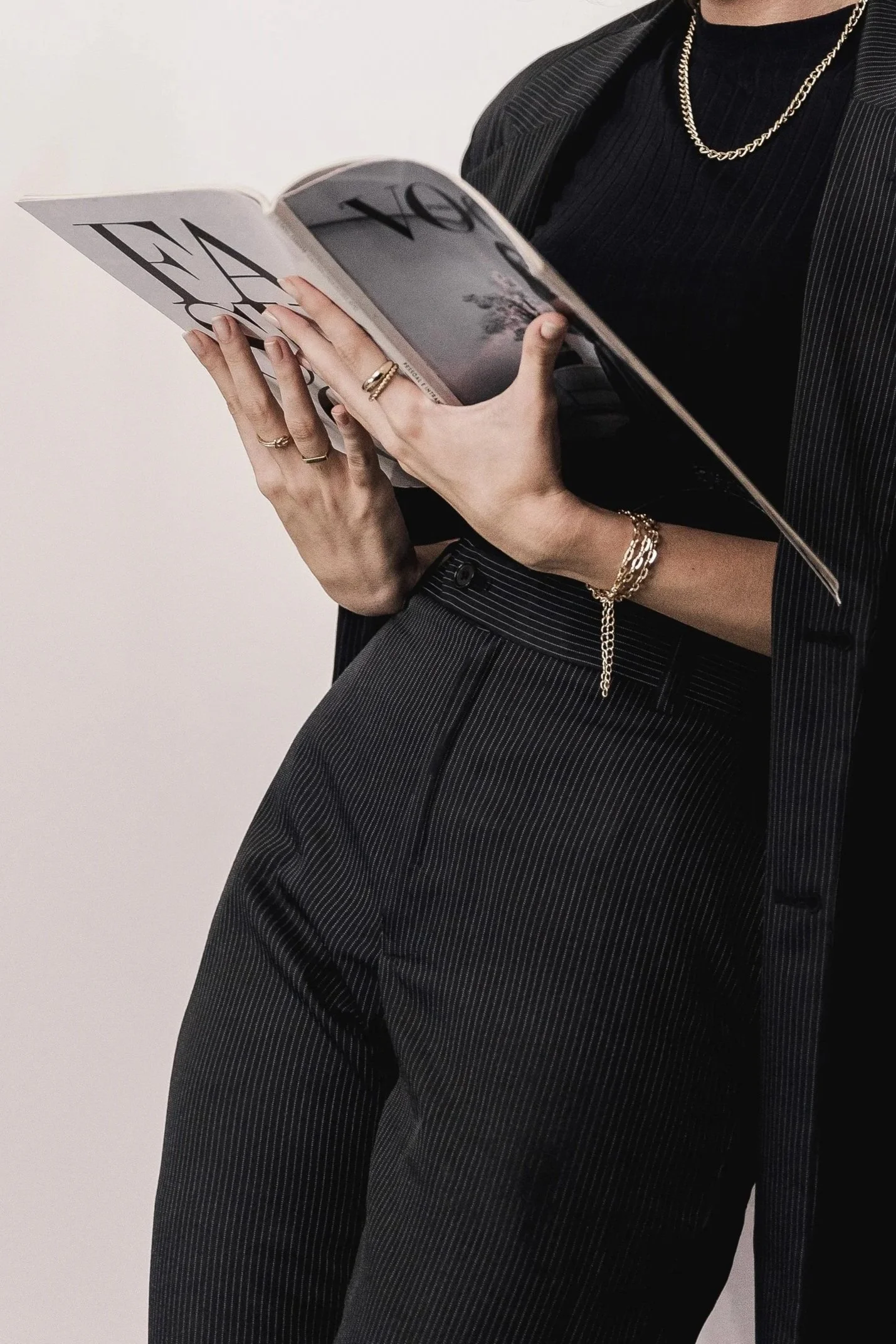 Person wearing black pinstripe suit holding a magazine, with jewelry including rings, bracelets, and necklaces visible.