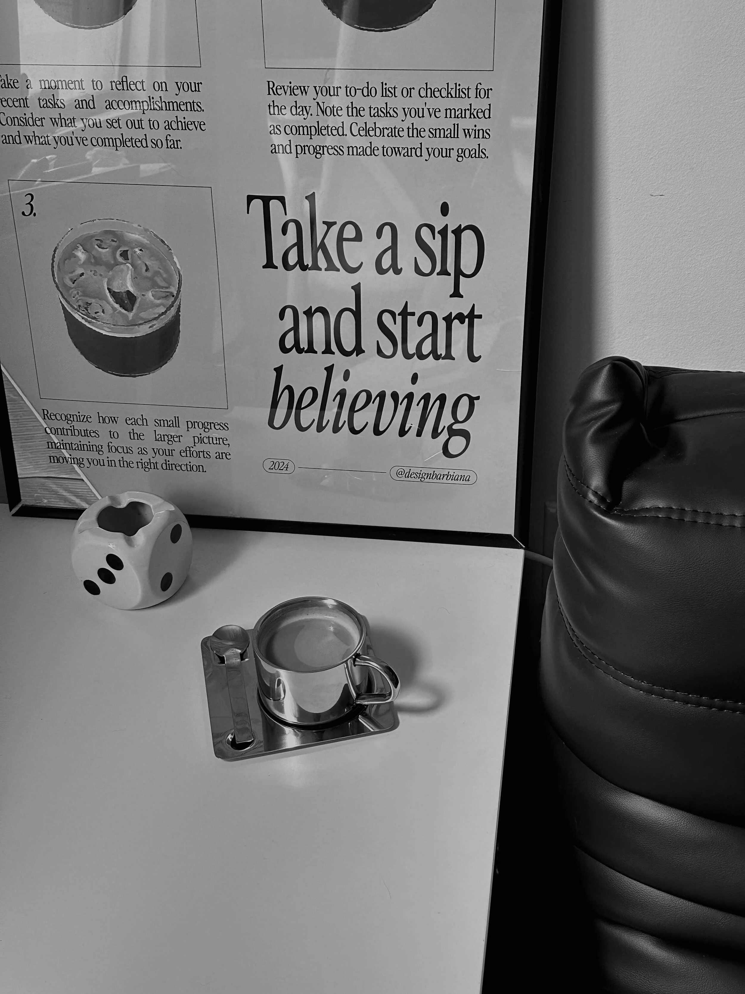 A small metal cup on a coaster, a ceramic dice-shaped container, and a framed poster with the phrase "Take a sip and start believing."