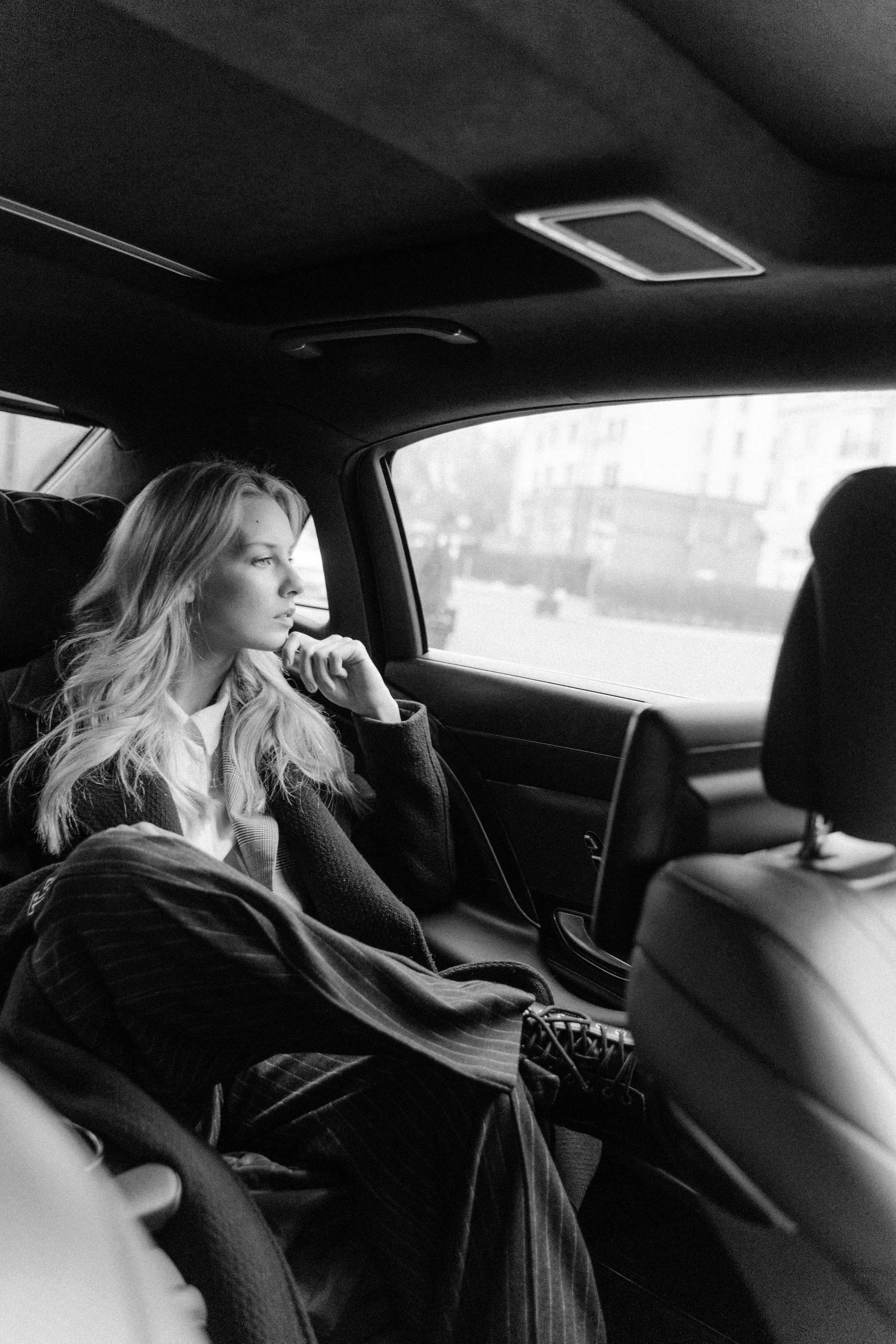 A young woman with long, wavy hair sitting in the back seat of a car, gazing out the window with a thoughtful expression, in black and white.