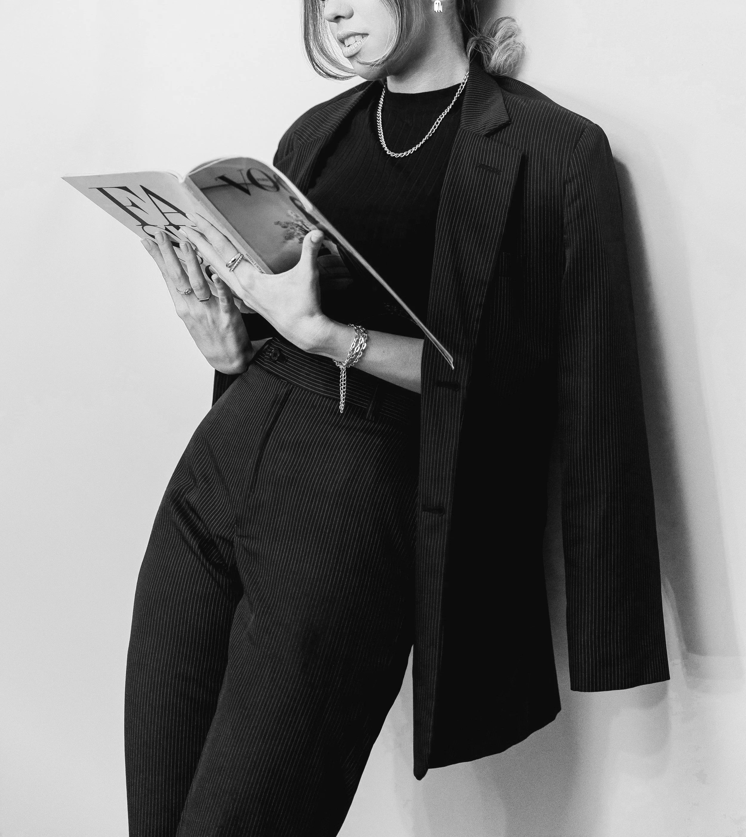 A woman in a pinstripe suit reading a magazine, standing against a plain wall, wearing jewelry including rings, a bracelet, a necklace, and a watch, in black and white.