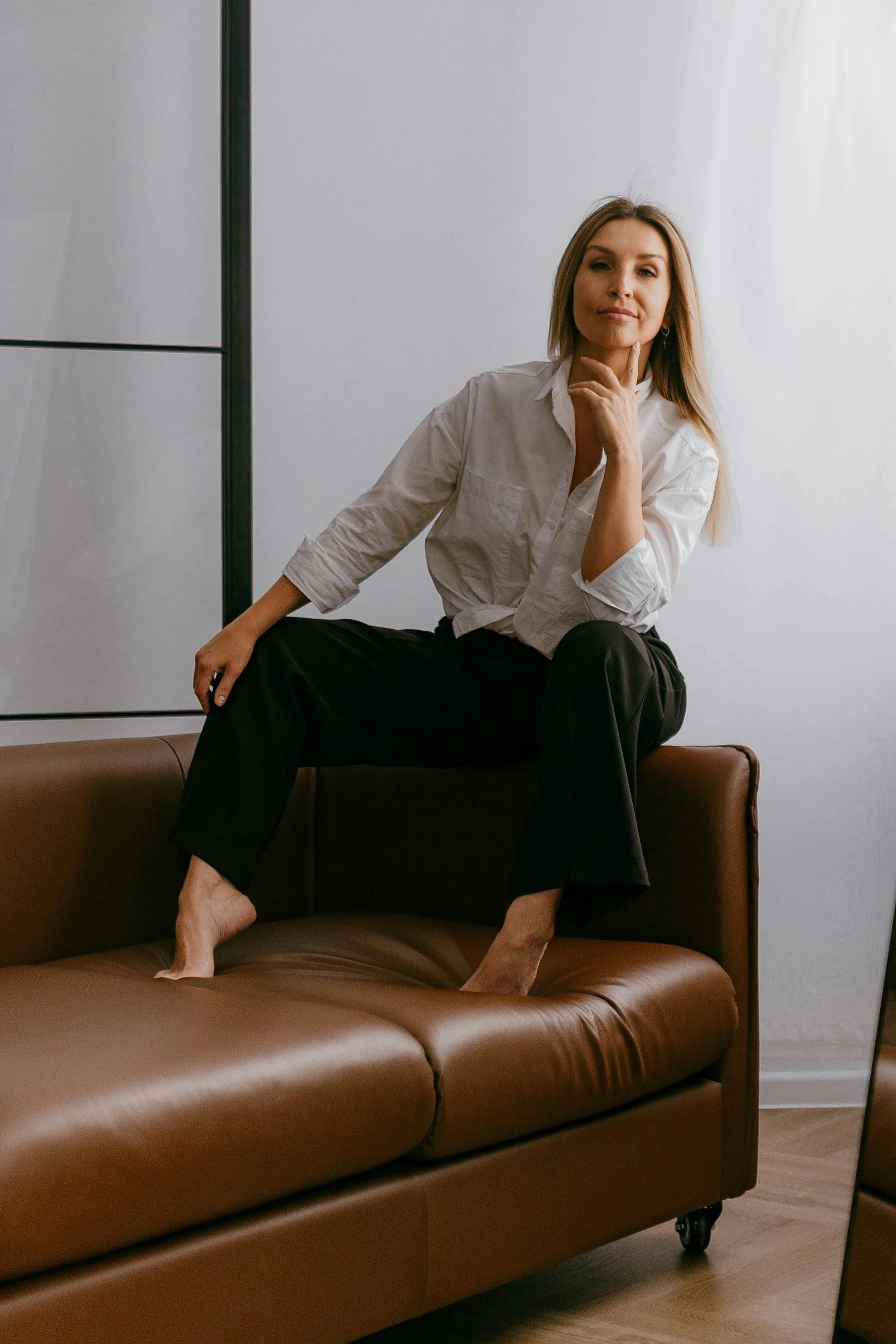 A woman sitting on a brown leather couch with one foot on the cushion and the other on the seat, dressed in a white shirt and black pants, with her hand resting on her chin, in a room with a gray wall and a mirror.