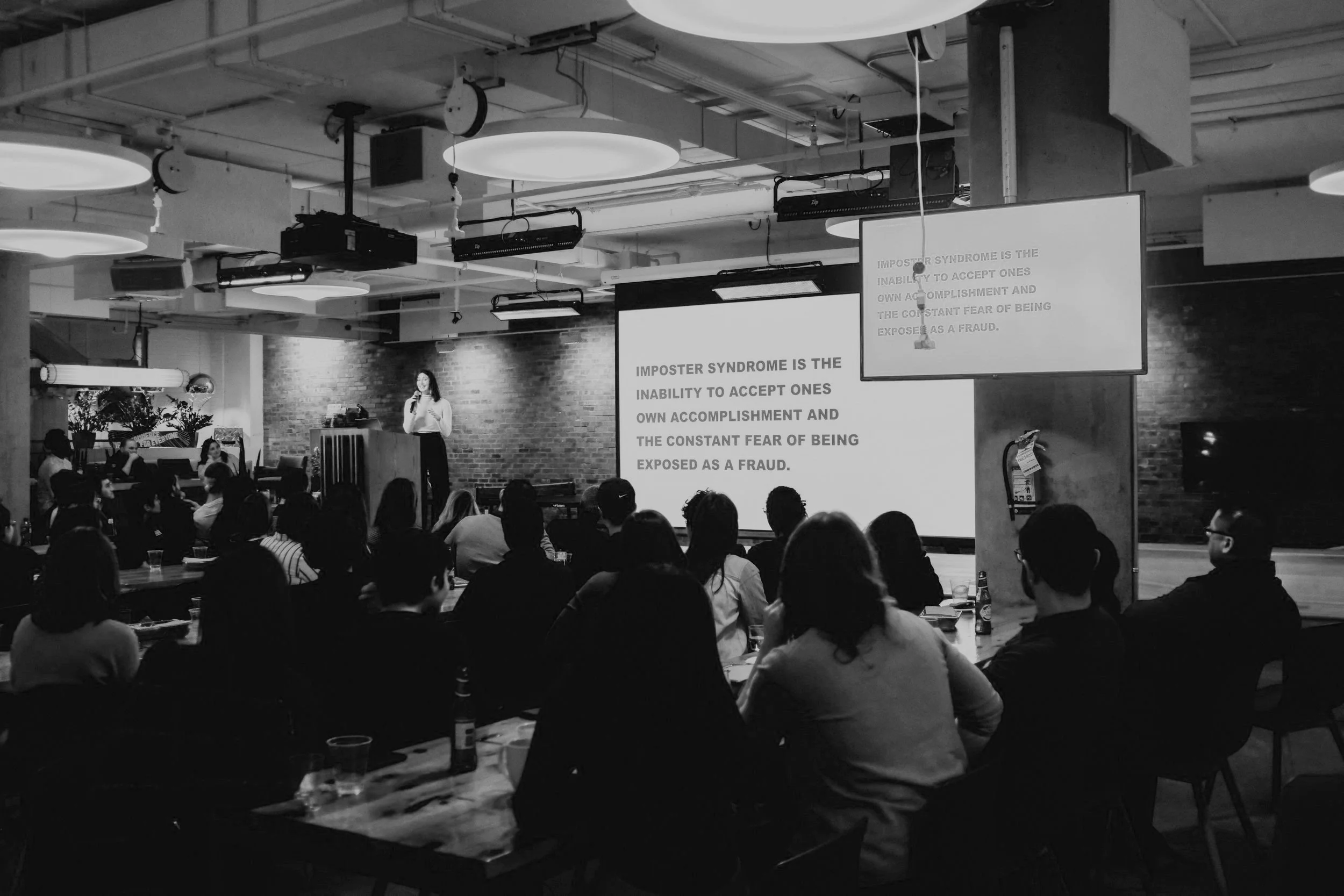 A woman giving a presentation in front of a large screen with a slide that reads, 'Imposter Syndrome is the inability to accept one's own accomplishment and the constant fear of being exposed as a fraud,' in a room full of seated audience members.