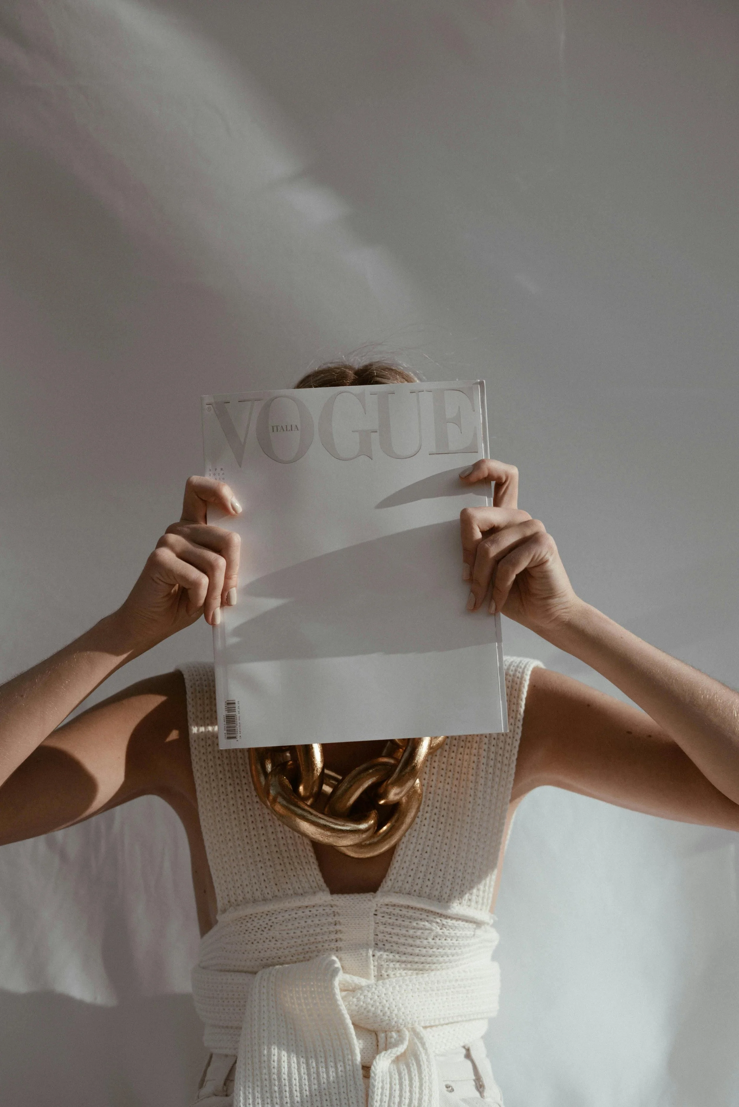 Person holding a Vogue magazine in front of their face, wearing a large gold chain necklace and a sleeveless cream-colored top with a tie at the waist. The lighting creates shadows on the wall and body.