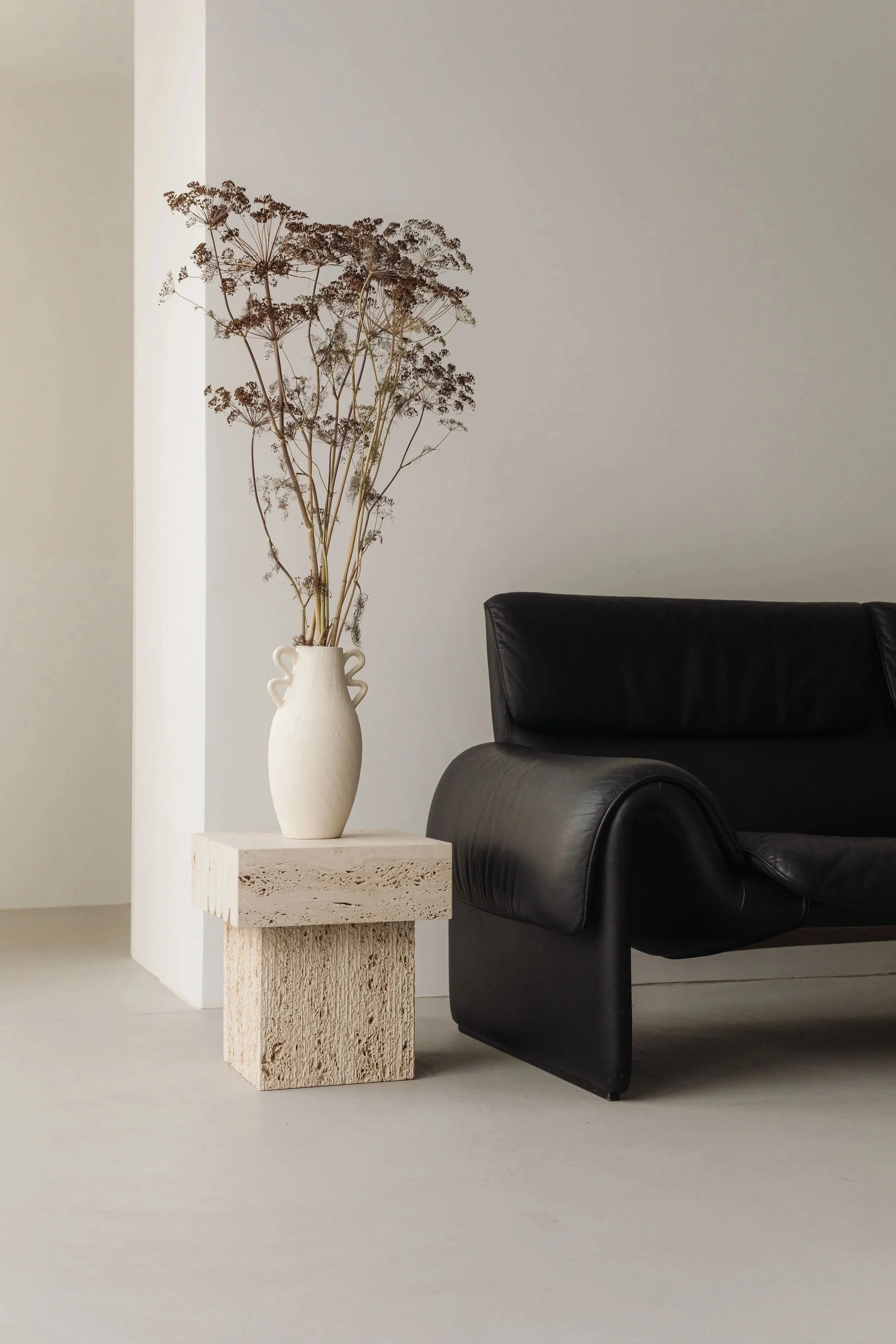 A minimalist living room corner with a black leather sofa, a textured white stone side table, and a white vase with dried branches, against a plain white wall.