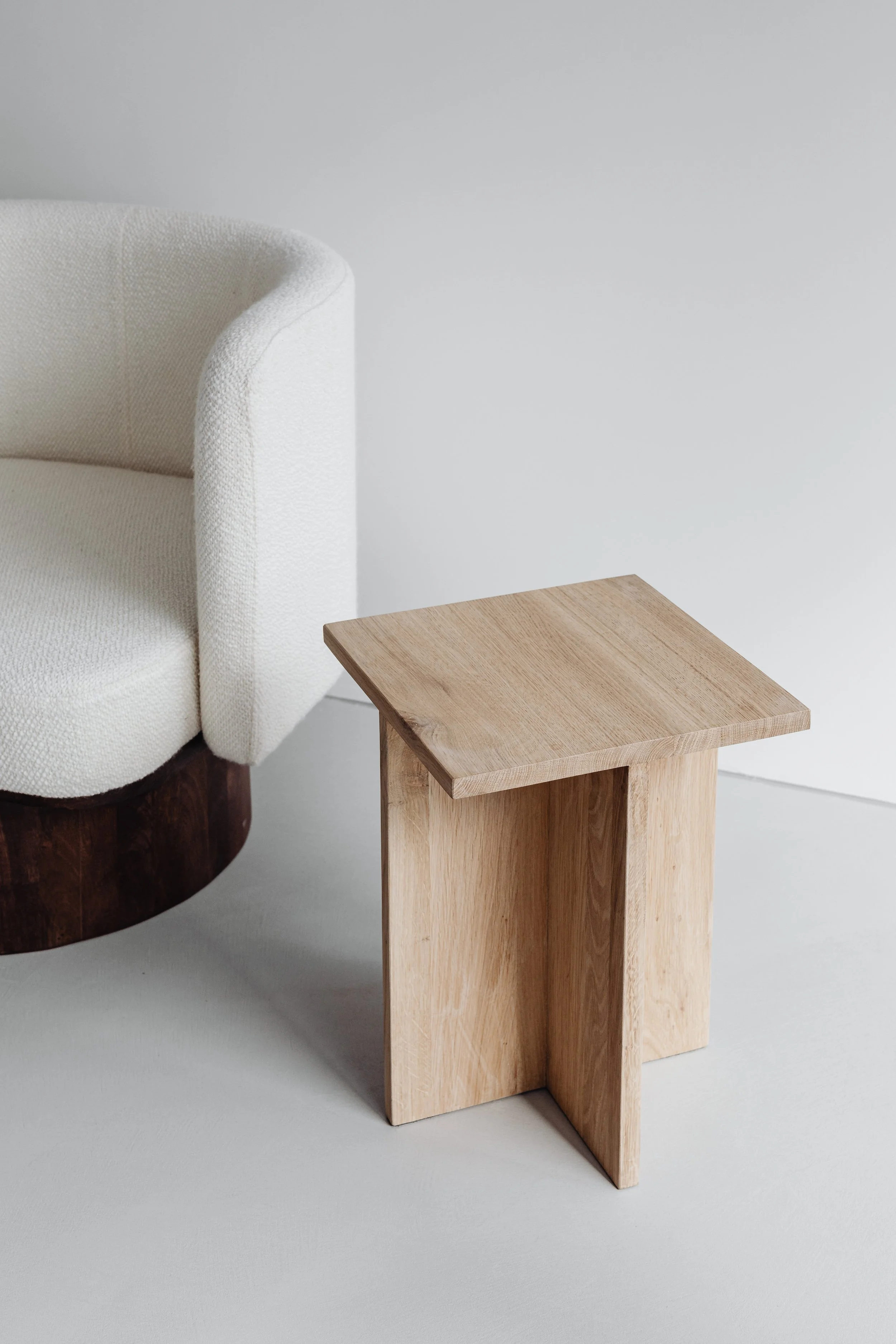 A wooden side table next to a cream-colored armchair on a white floor.