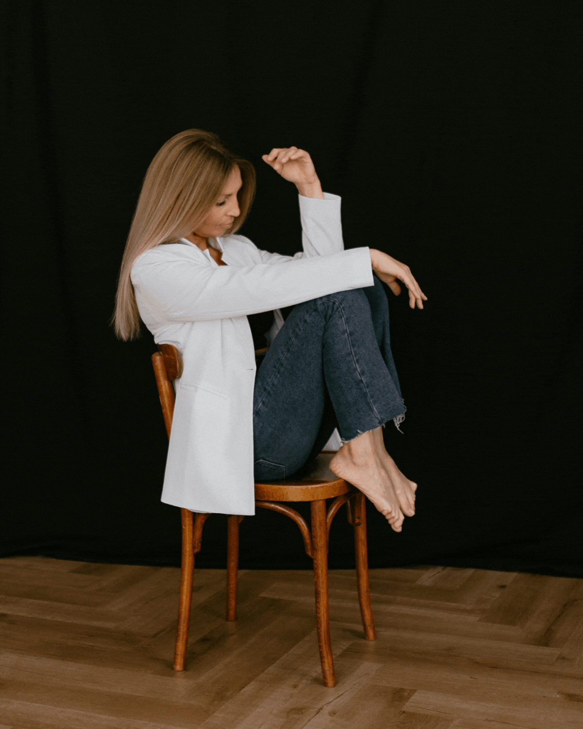 A woman sitting on a wooden chair with her knees drawn to her chest against a black background. She has long, straight, blonde hair, is wearing a white blazer and dark jeans, and is barefoot. She is looking down with her eyes closed and has one arm raised, bent at the elbow.
