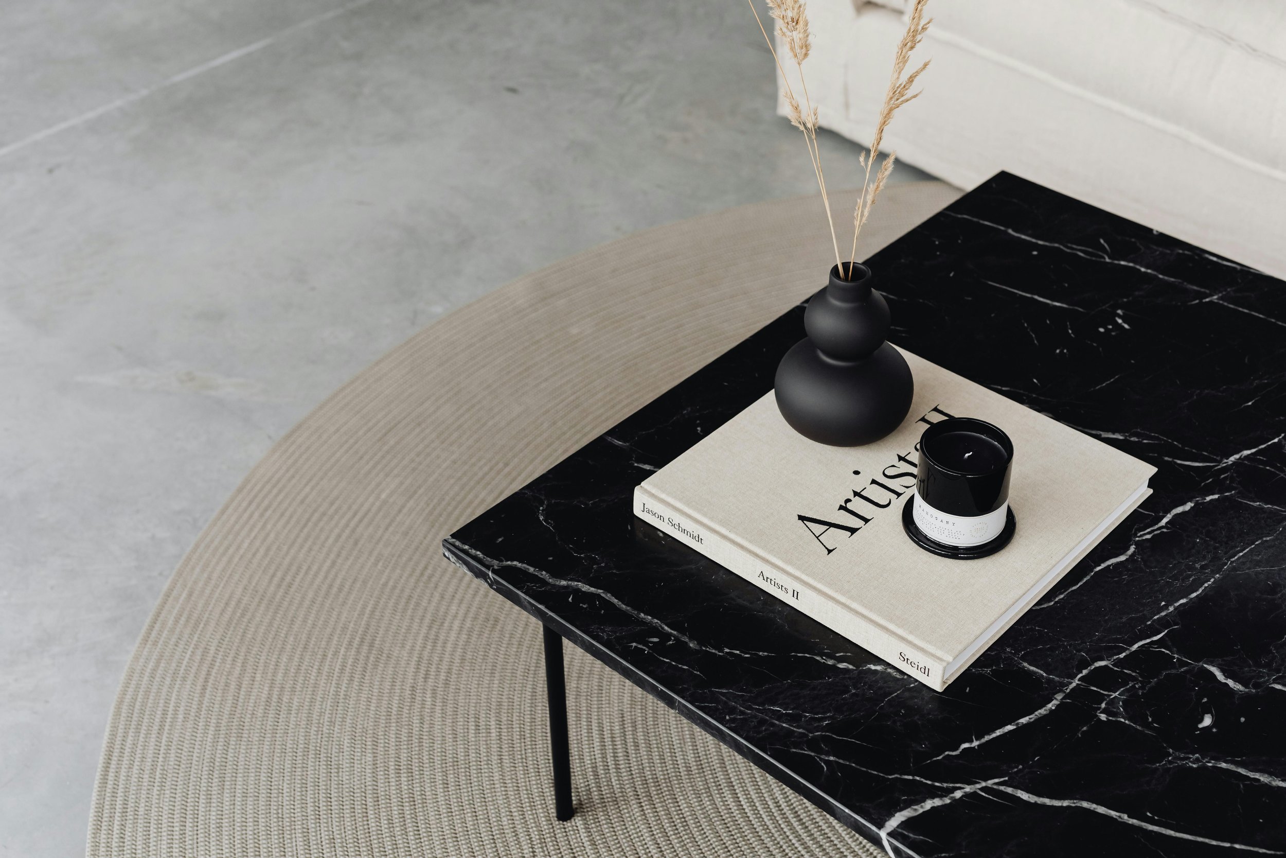 A black marble table with white veining, on top of a beige round rug. The table holds a beige book titled "Artists" by Jason Schmidt, a black candle in a white holder, and a black matte vase with tall dried grass stems.