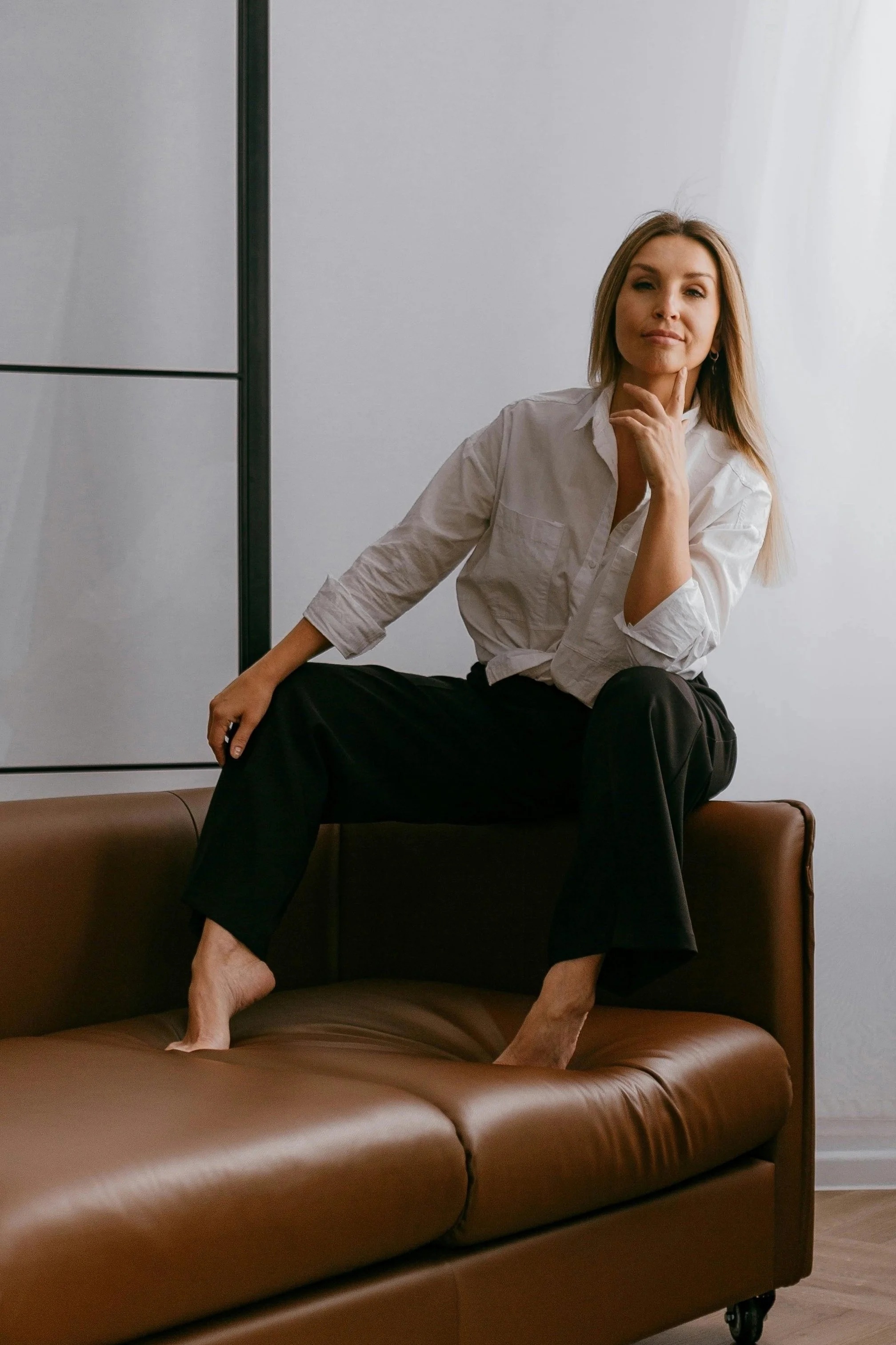 A woman with blonde hair, wearing a white shirt and black pants, sitting on a brown leather sofa in a relaxed pose with one leg up and hand on her chin, next to a minimalist wall.