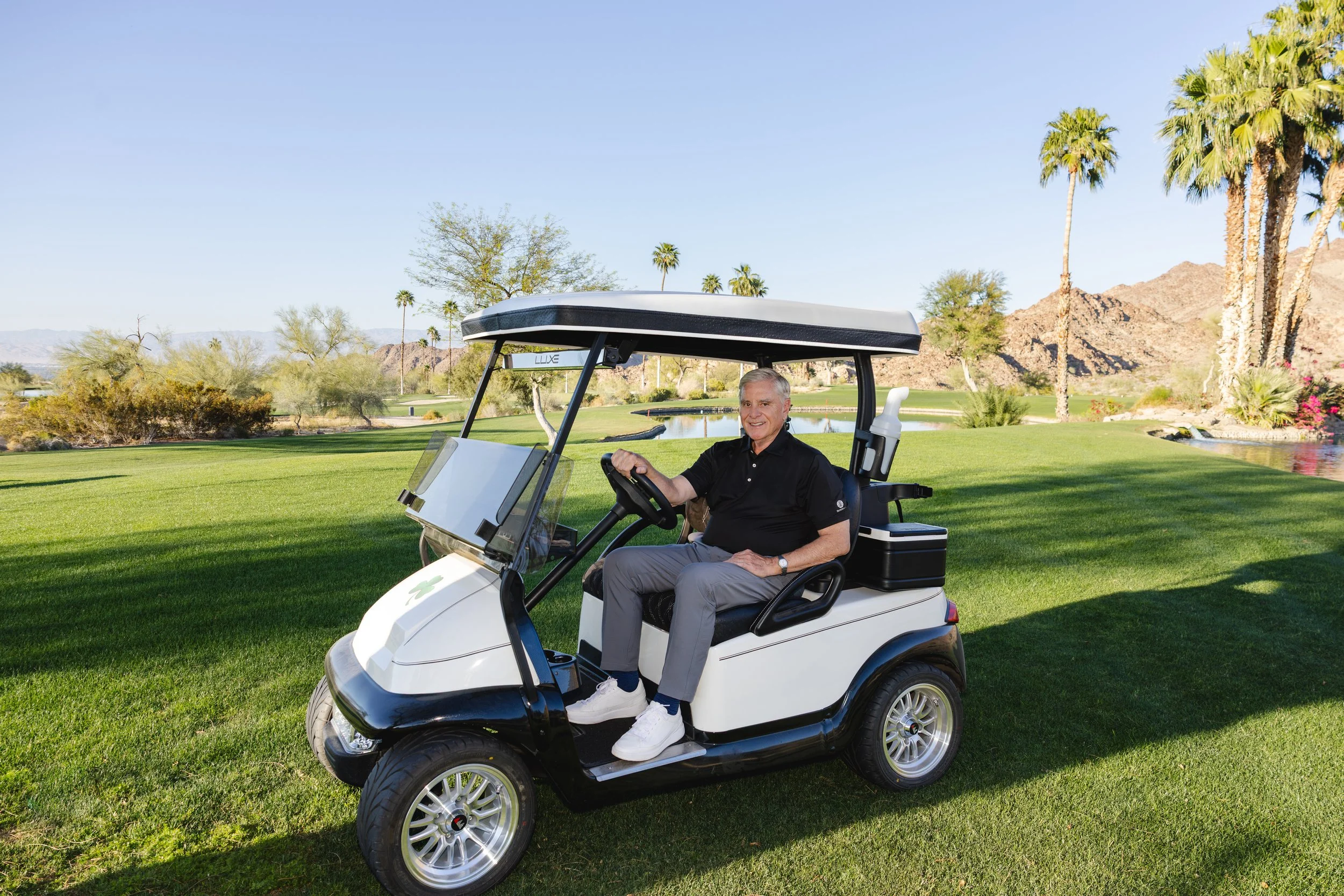 A man sitting in a golf cart on a golf course with green grass, palm trees, mountains, and a clear blue sky in the background.