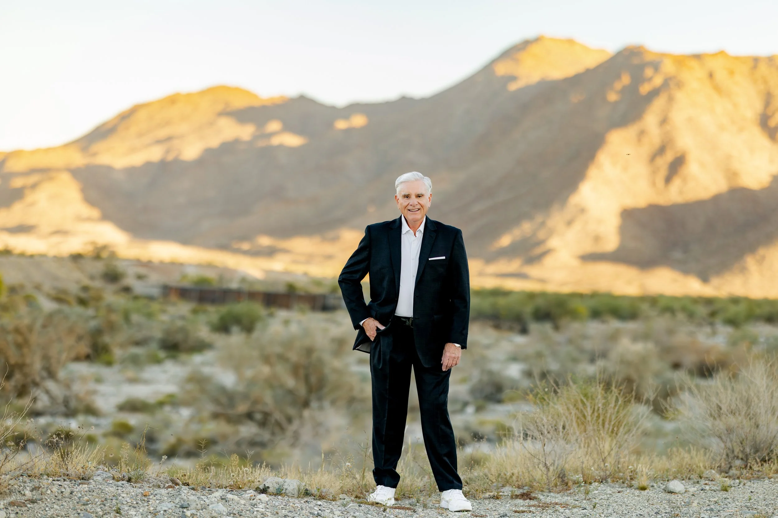 An elderly man in a black suit and white sneakers standing outdoors in a desert landscape with mountains in the background.