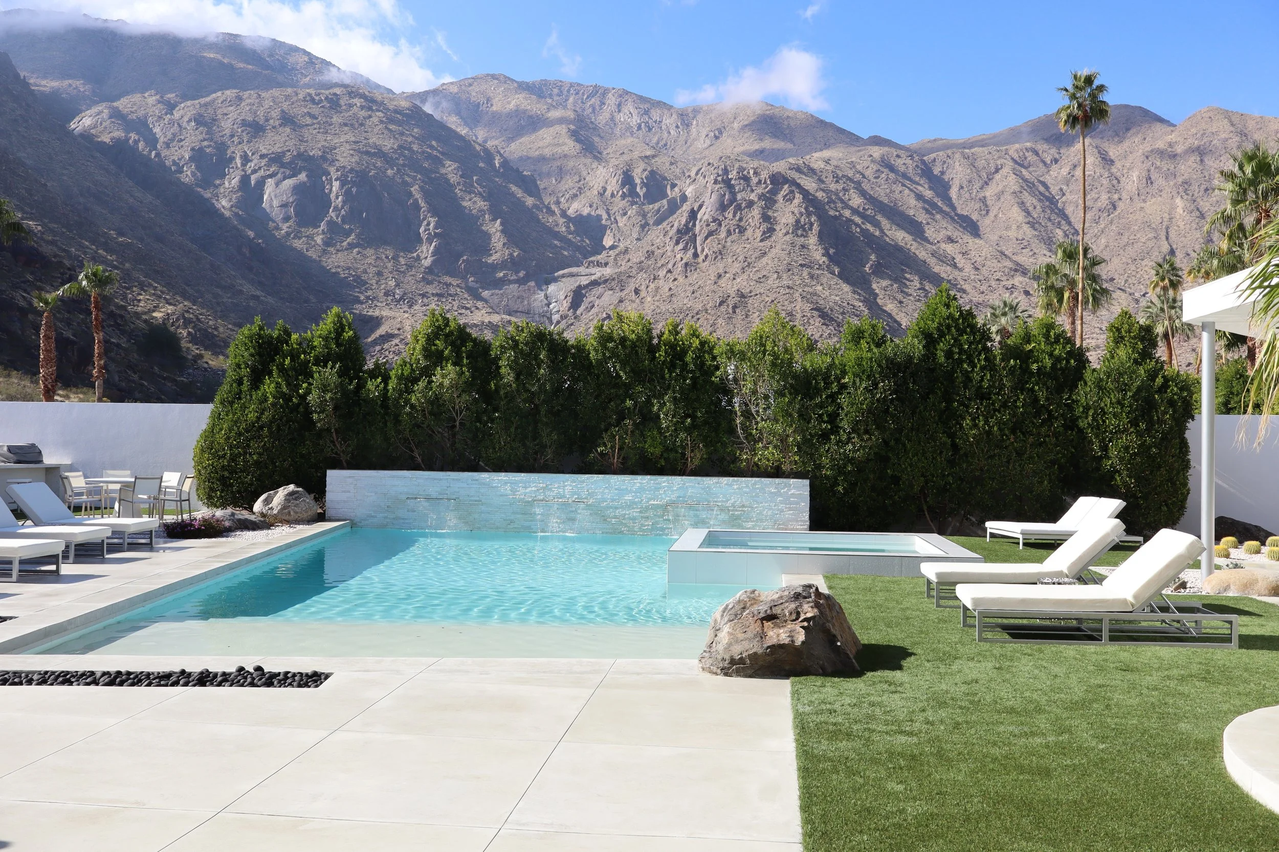 Luxury backyard with a modern rectangular swimming pool, lounge chairs, lush green bushes, desert mountain backdrop, and clear blue sky.