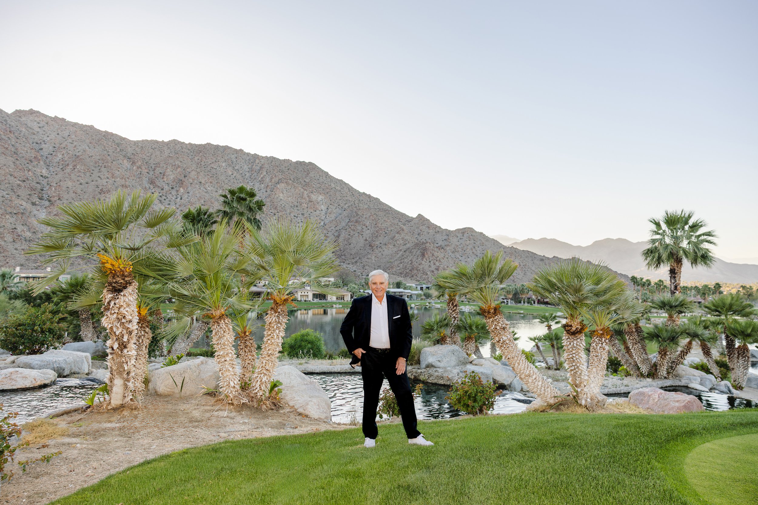 A man in a black suit and white shirt standing on a manicured lawn with palm trees, rocks, and a lake, mountain landscape in the background.