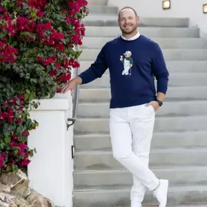 A man with a beard wearing a blue sweater with a bear design, white pants, and white shoes, standing on stairs next to a bush with pink flowers, smiling.