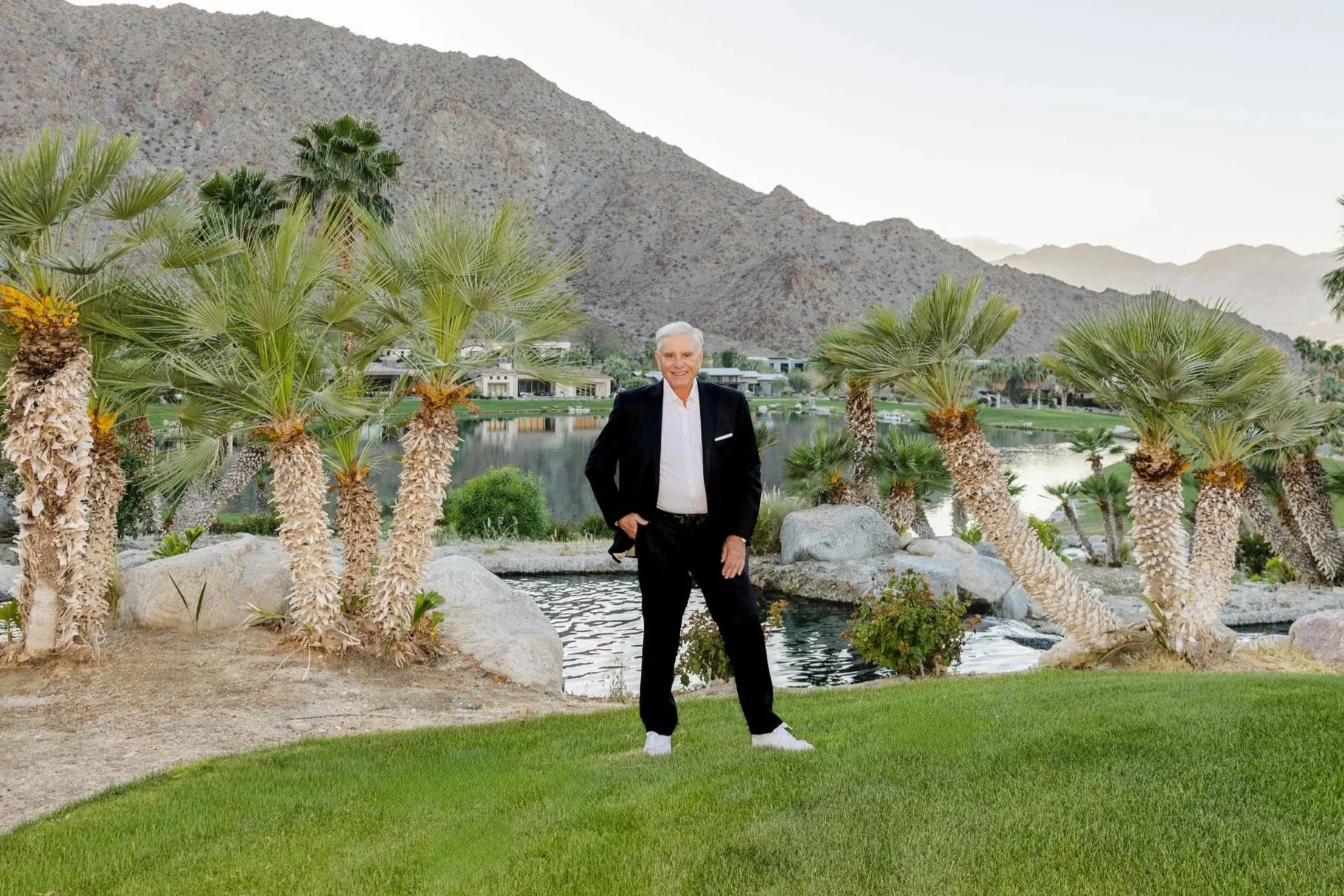 A man in a black suit and white shirt standing on grass near a pond with palm trees and mountains in the background.