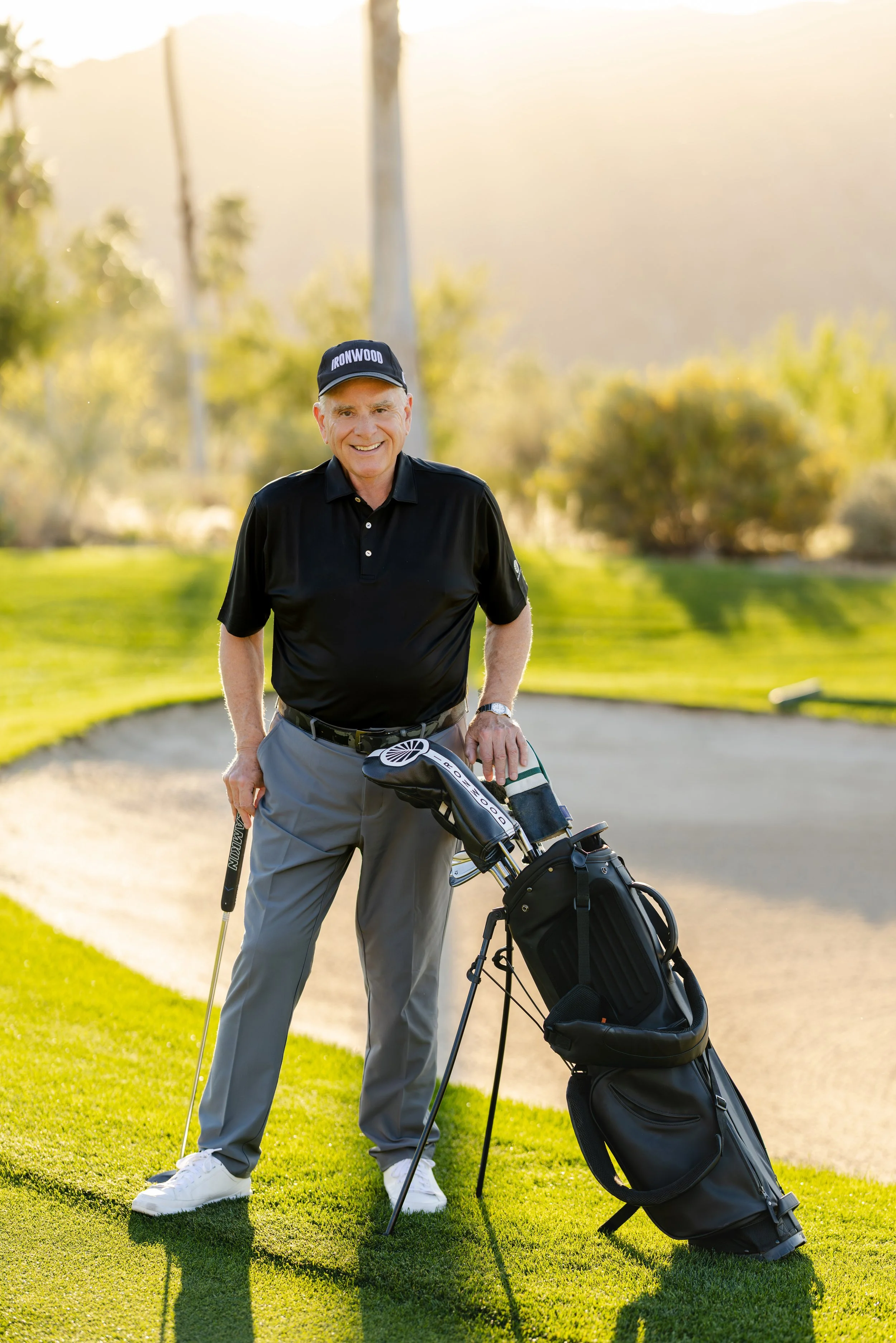 A man standing on a golf course with a golf club and a golf bag, smiling at the camera during sunset.