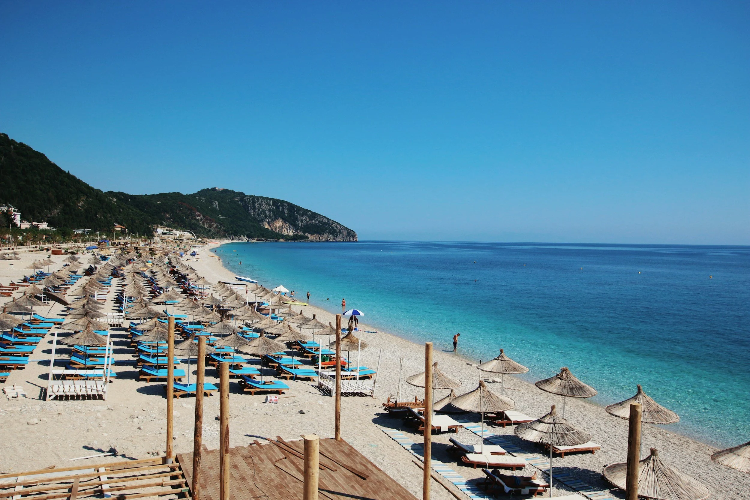 Quiet sandy beach with arranged sun loungers and straw umbrellas, clear blue water, green hillside, and few people walking or relaxing.