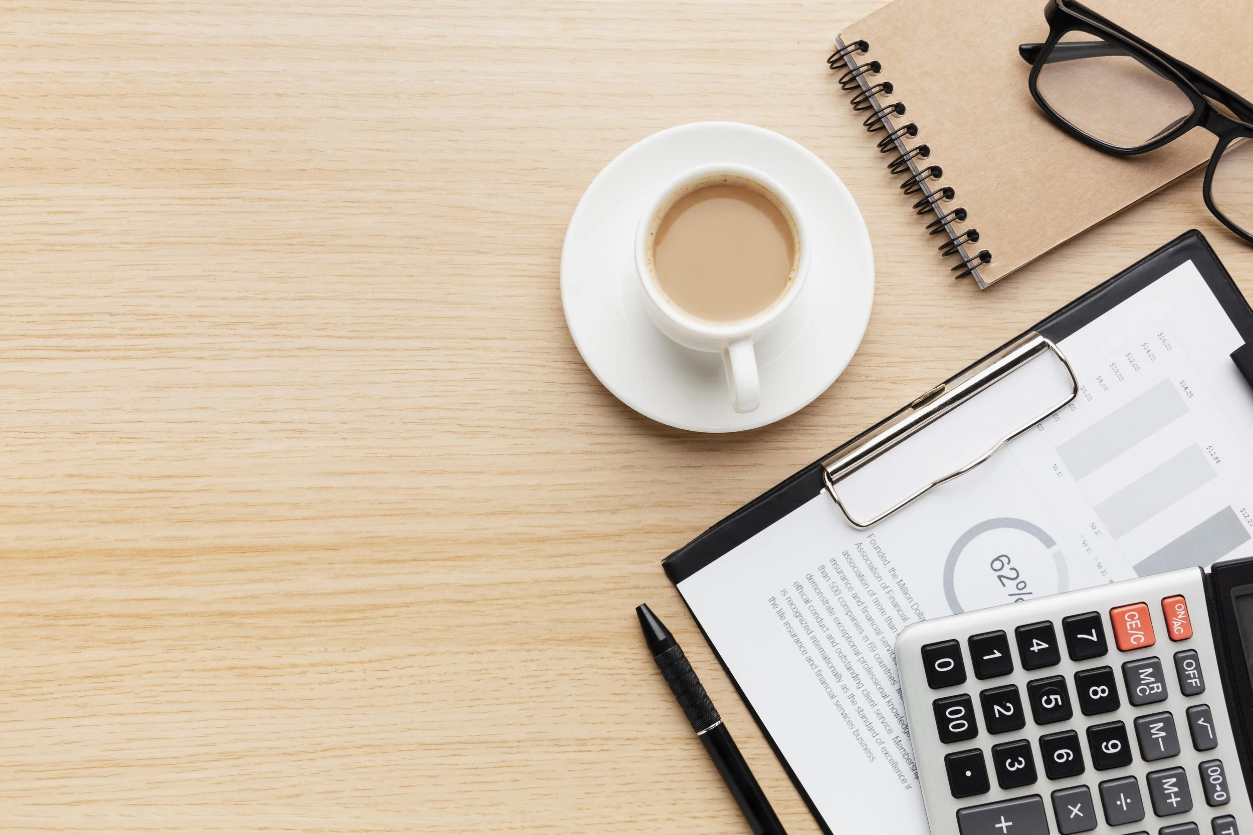 Work desk with a cup of coffee on a white saucer, a clipboard with a document and calculator, a black pen, a notebook, and a pair of glasses on a light wood surface.