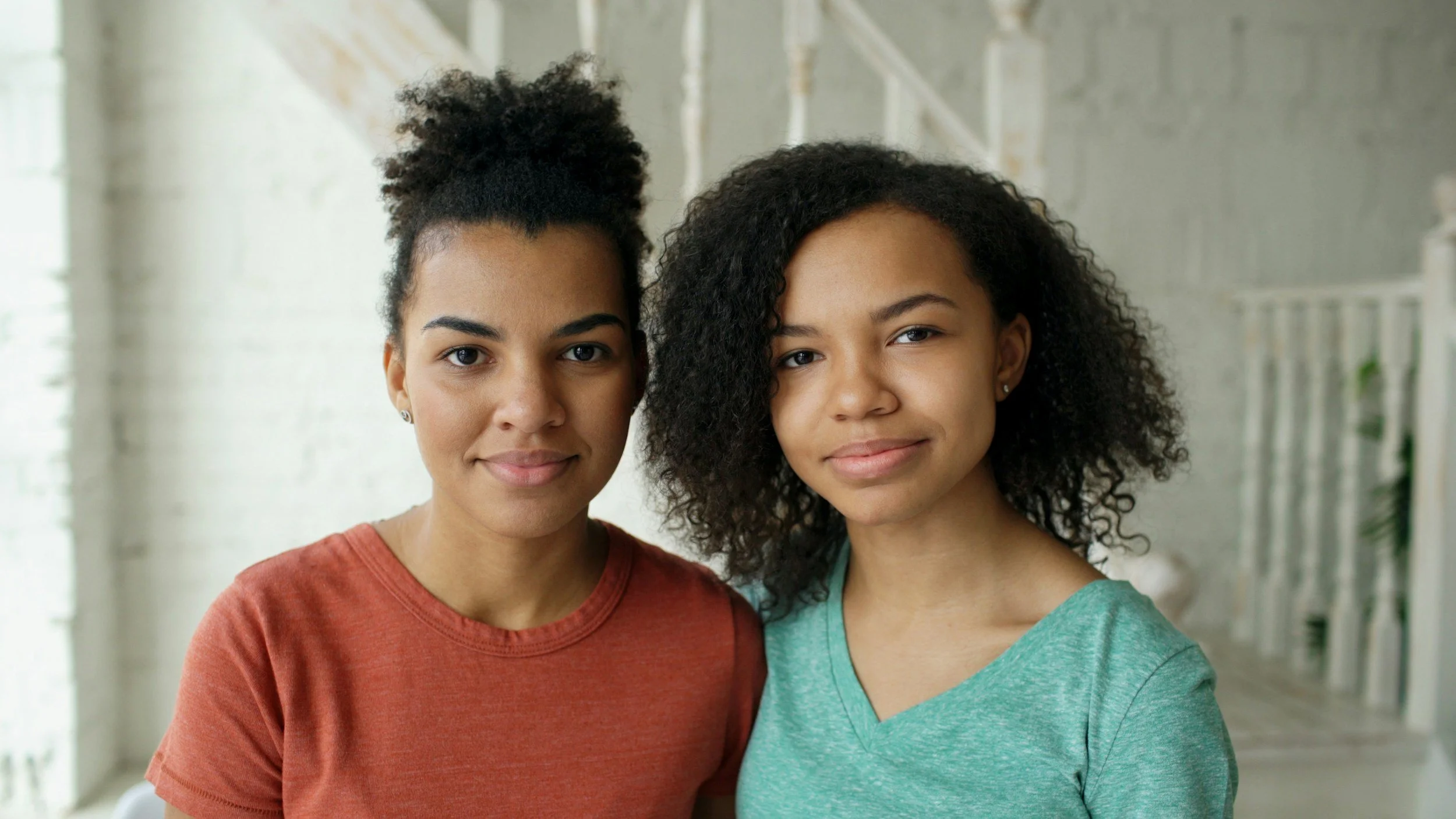 Two young women with curly hair stand close to each other indoors, smiling softly at the camera.