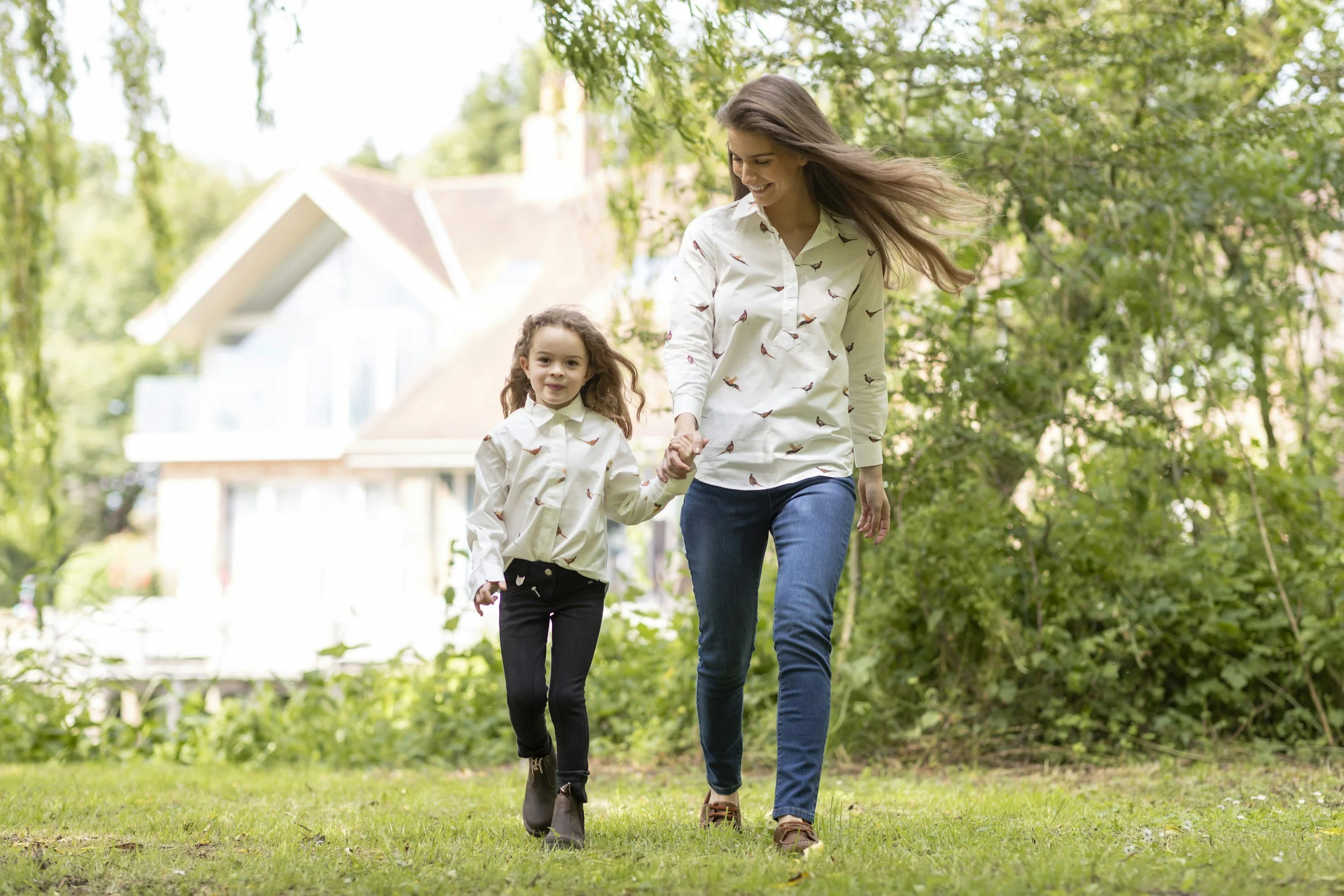 A woman and a young girl are walking hand in hand outdoors on a grassy area, surrounded by trees with a house visible in the background. Both are wearing matching white shirts with a bird pattern.