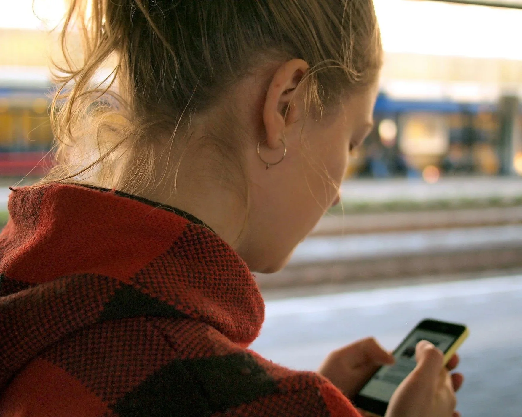 A woman with braided blonde hair wearing a red and black plaid shirt and hoop earrings is looking at her smartphone at a train station during daylight.