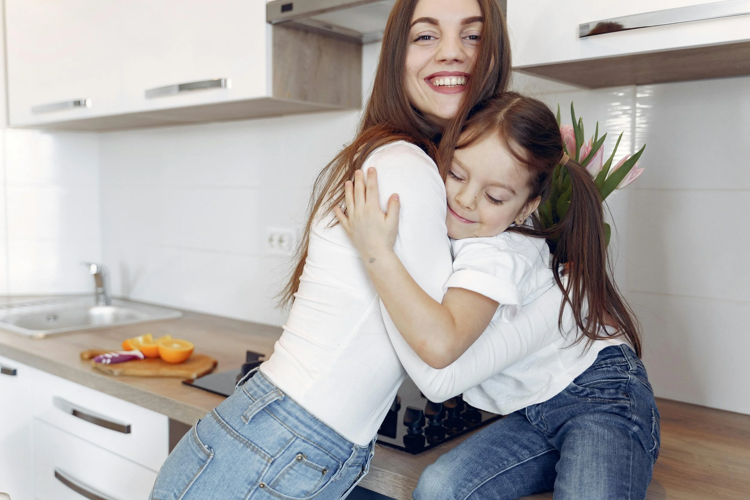 A woman and a girl hugging in a modern kitchen, smiling with the girl holding a bouquet of pink tulips.