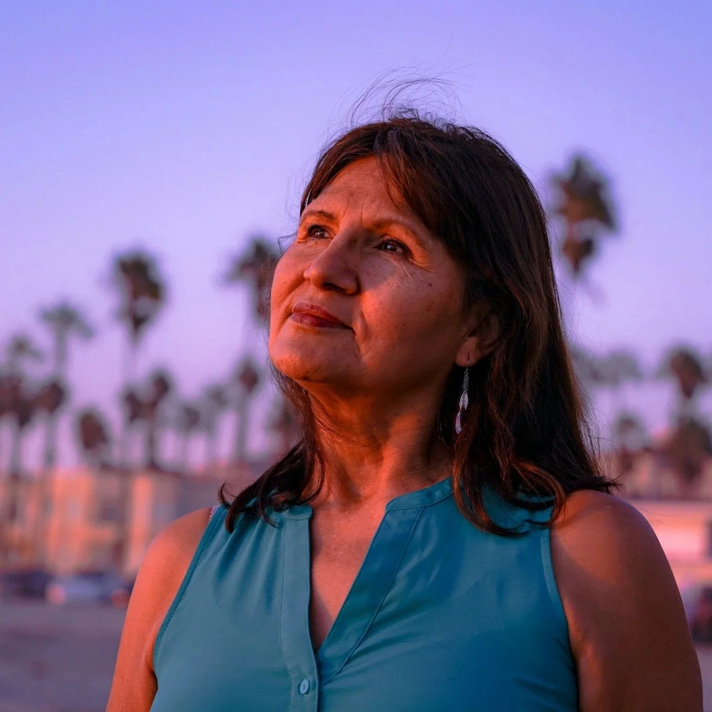 A woman with dark hair looking thoughtfully into the distance with palm trees in the background at sunset.
