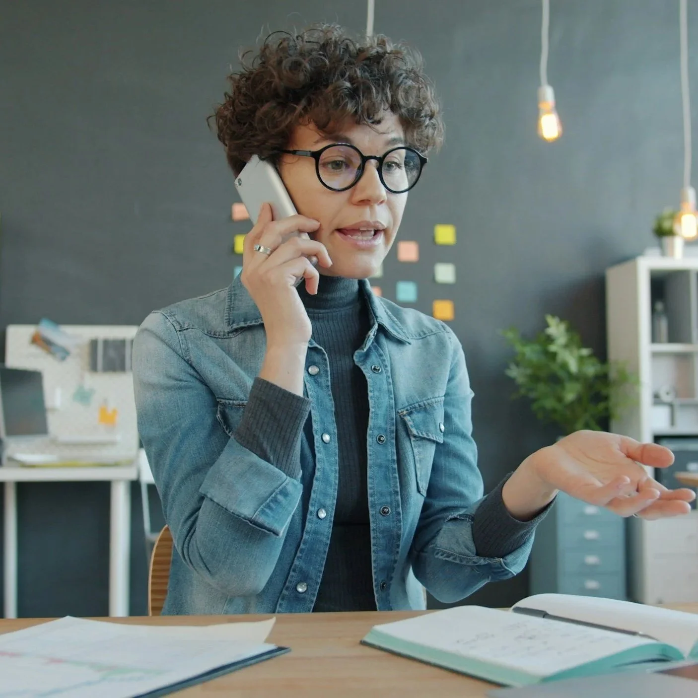 A woman with curly hair and glasses, wearing a denim jacket and turtleneck, talking on the phone and gesturing with her other hand in an office setting with papers and a notebook on the desk.