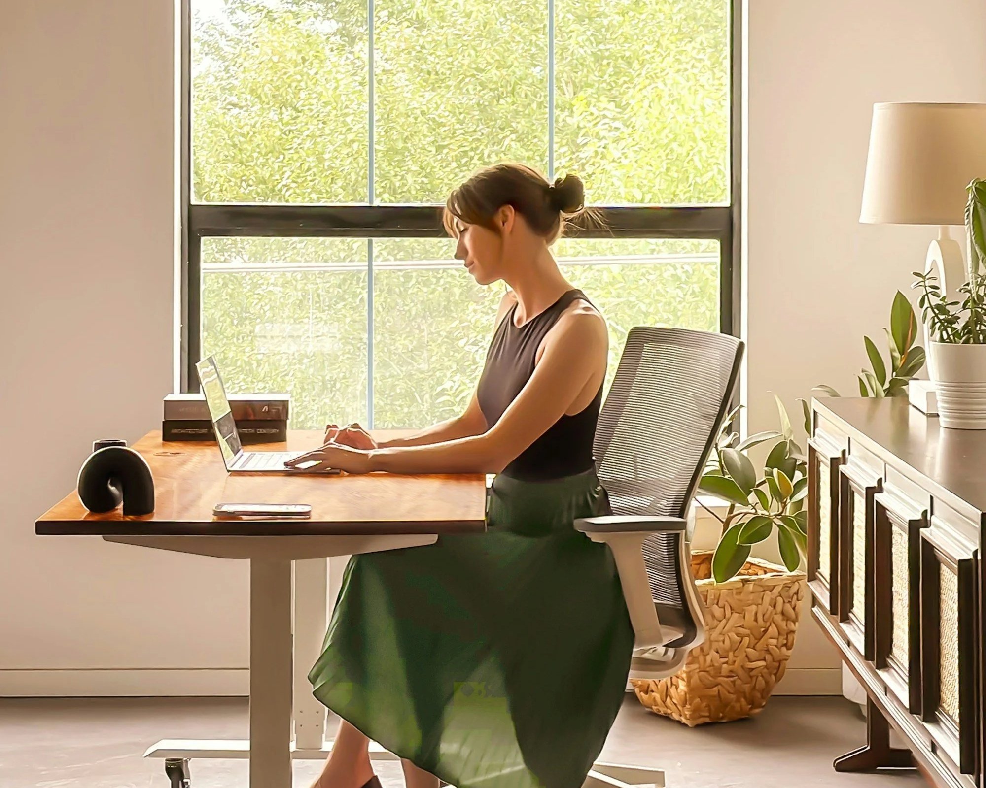 A woman working on a laptop at a wooden desk in a bright, modern office with large windows and houseplants.