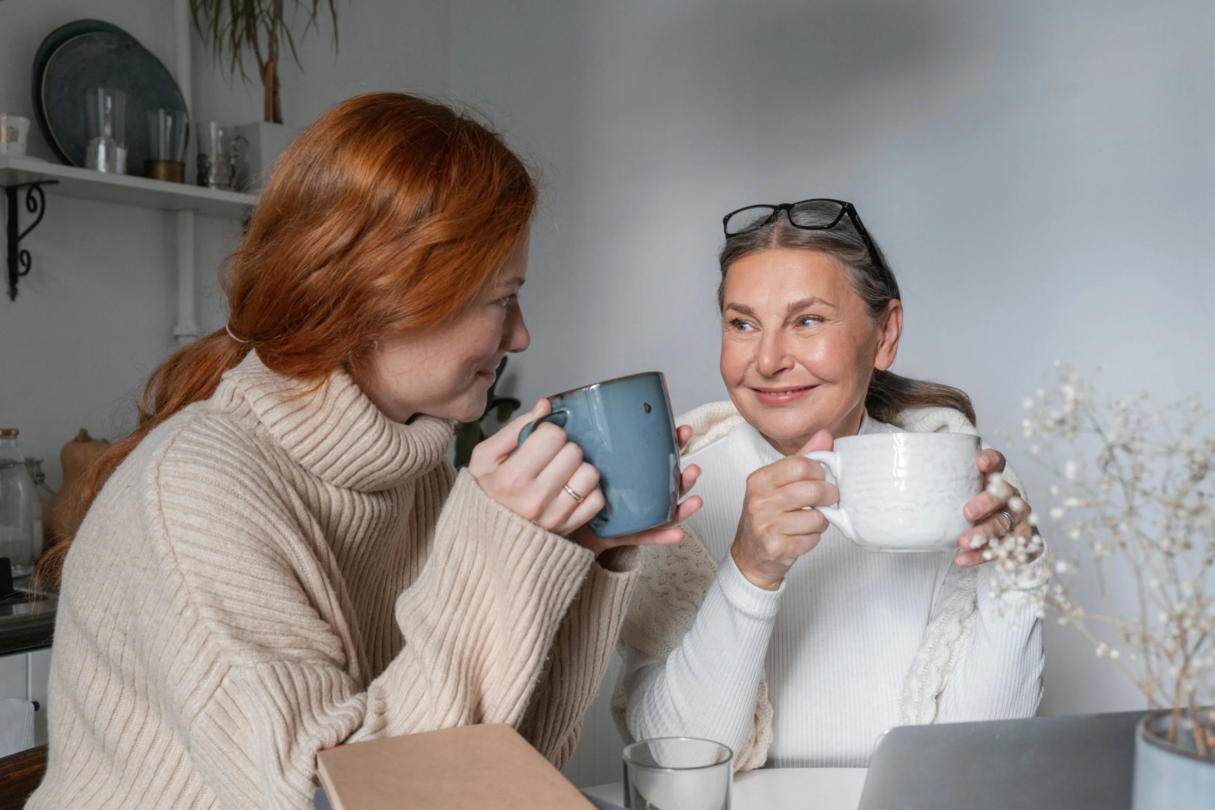 Two women, one with red hair and the other with gray hair, sit at a table holding mugs and smiling at each other in a cozy domestic setting.