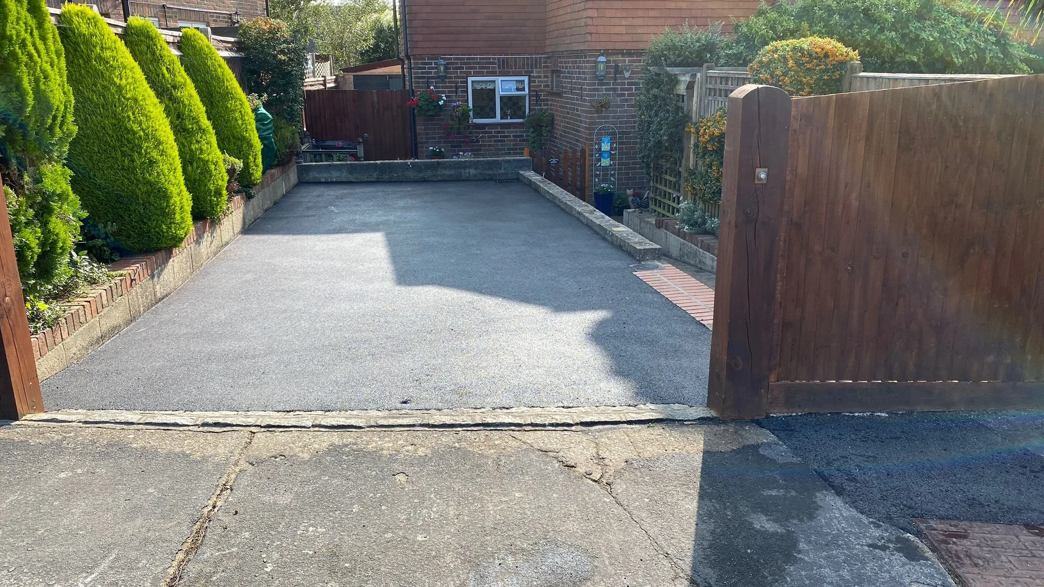 View of a small paved backyard with a wooden gate on the right side and a brick wall at the back, with potted plants and shrubs along the sides.