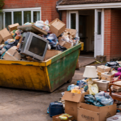 Large green dumpster filled with trash, old electronics, and cardboard boxes outside a house. Additional trash and boxes surrounding it on the ground.