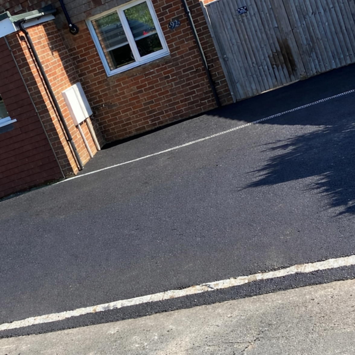 Photo of a house with a brick wall, window, and a wooden fence, along with a newly paved parking space marked with white lines.