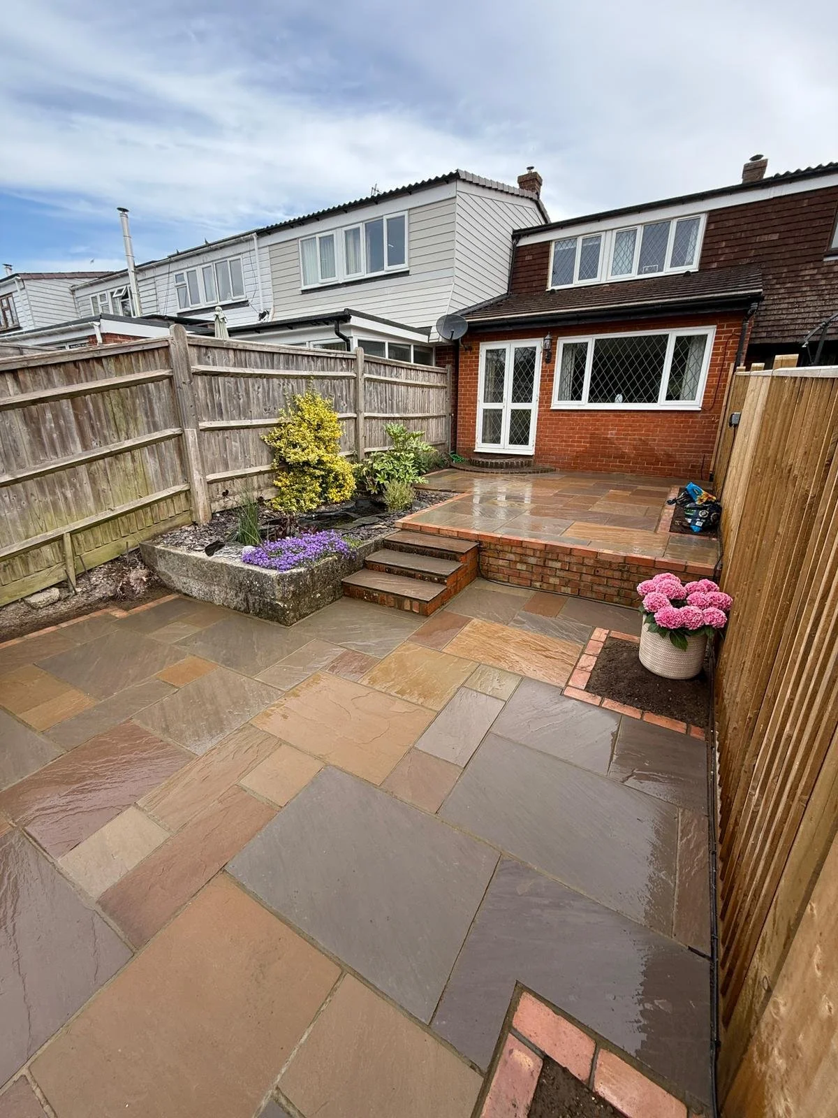 A backyard patio with wet tile flooring, a small step leading to a higher level with a brick border, a garden bed with purple and yellow flowers, pink hydrangeas in a pot, wooden fences on both sides, and a brick and white house with windows in the background.