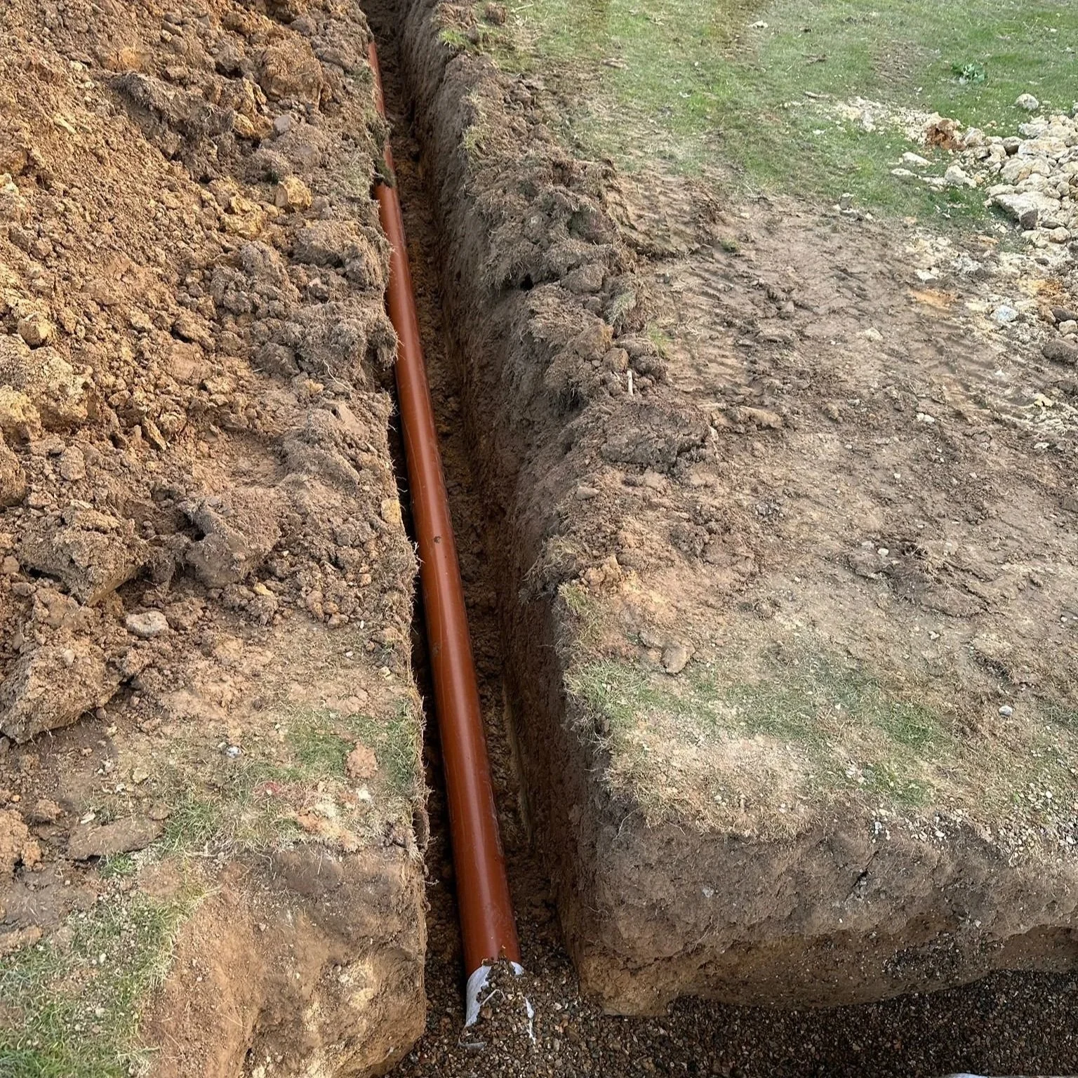 A new orange underground pipe runs through a narrow trench in a dirt and grass area, with loose soil piled on either side of the trench.