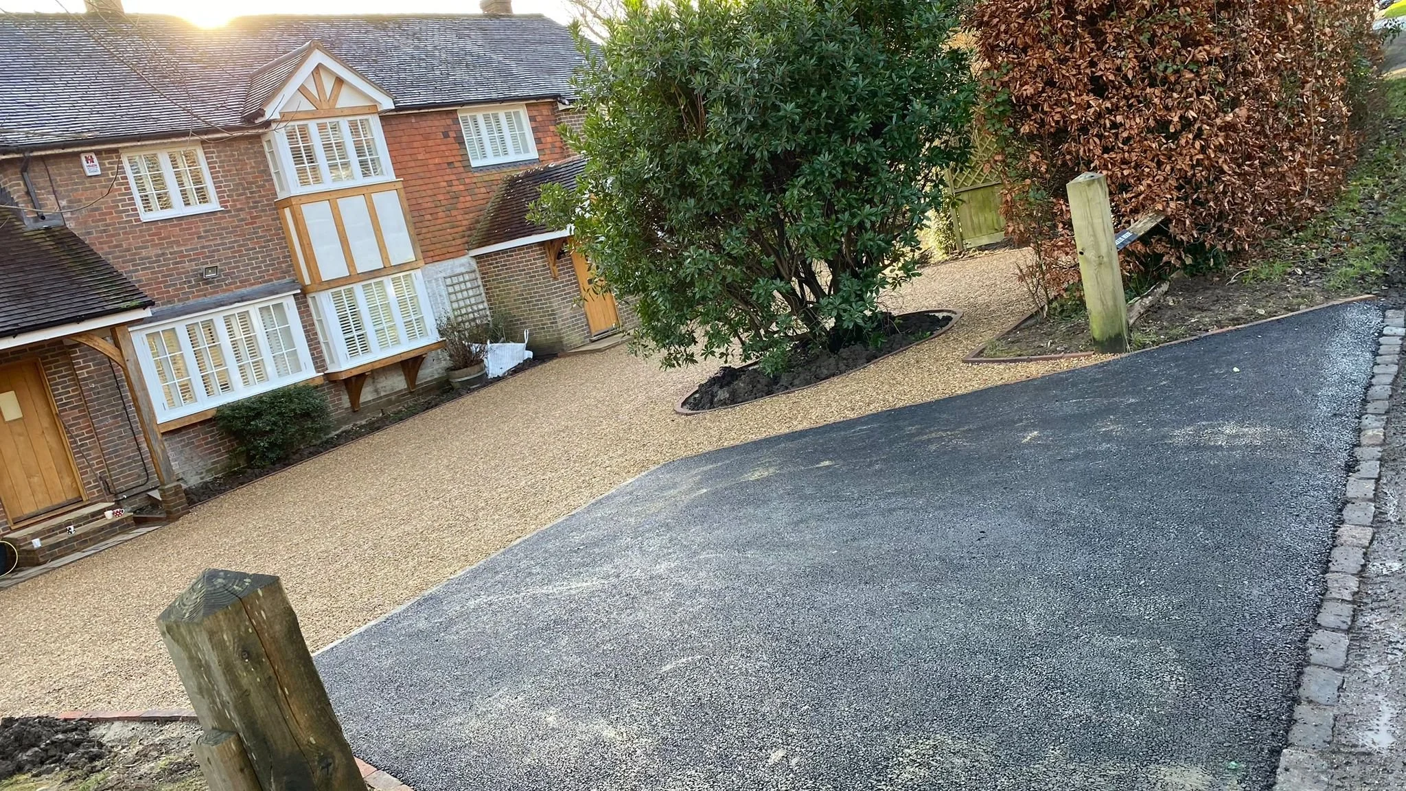 A newly paved driveway and gravel pathway in front of a brick house with white window shutters and a front door, surrounded by trees and a garden.
