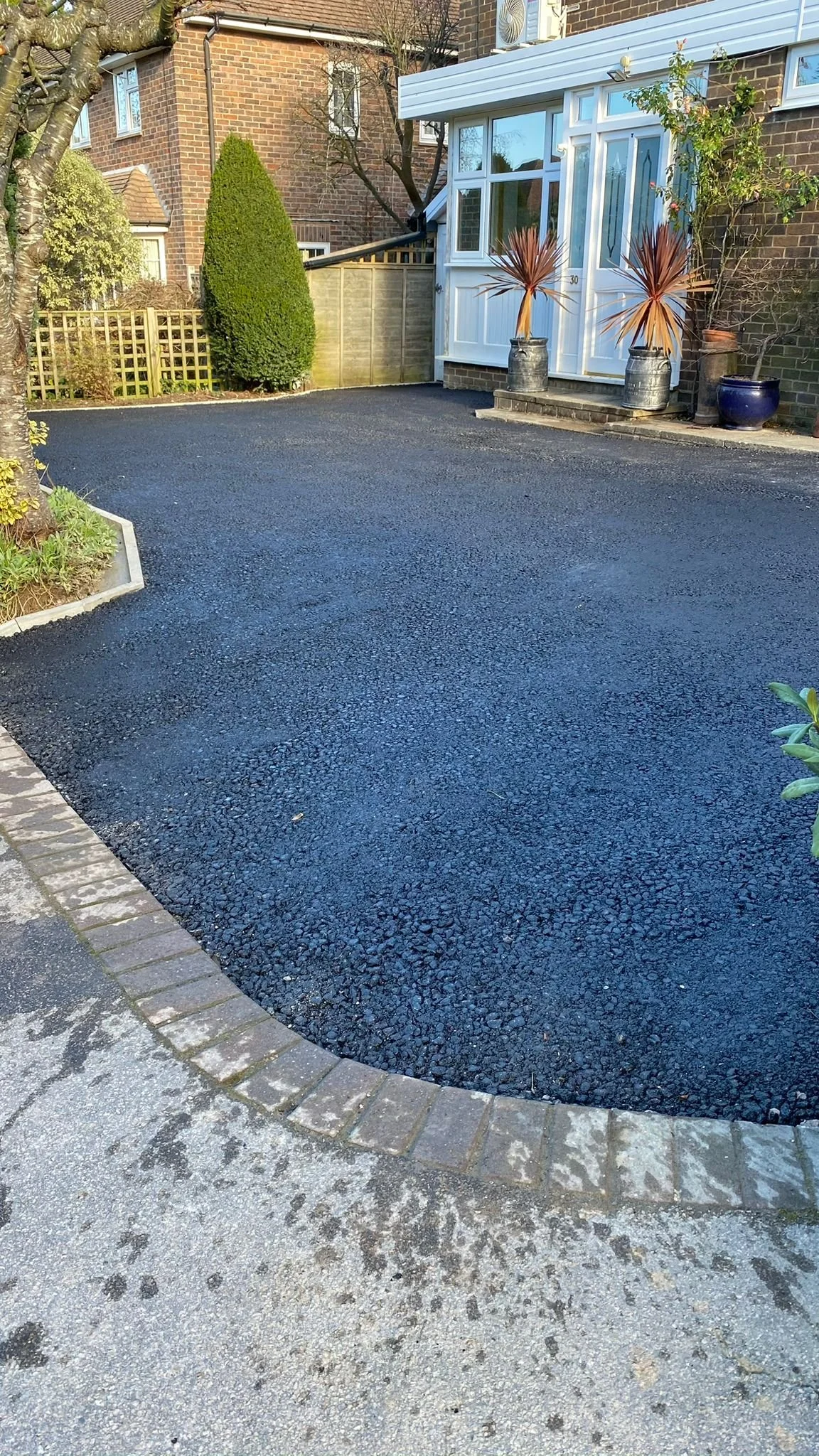 A newly paved black asphalt driveway in a residential backyard, bordered by a brick and concrete curb, with a few plants and trees in the background.