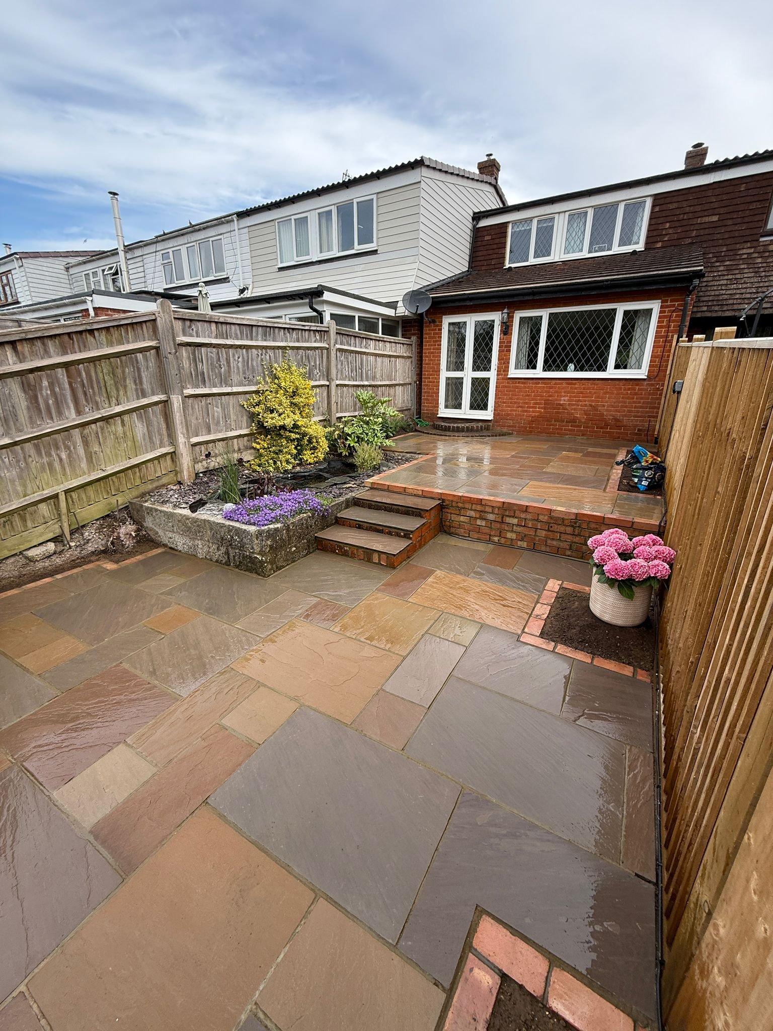 Backyard with wet stone patio, wooden fence, small garden bed with purple flowers and yellow-green shrub, brick steps, pot of pink flowers, and an aluminum door leading to a red brick house with white-framed windows.