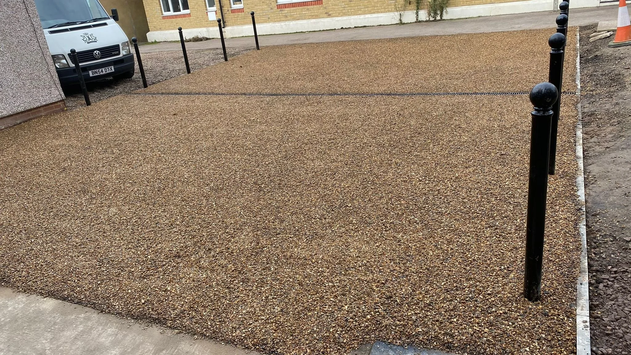 Newly installed gravel parking space with black metal posts as boundary markers. Part of a brick building and a white van are visible in the background.