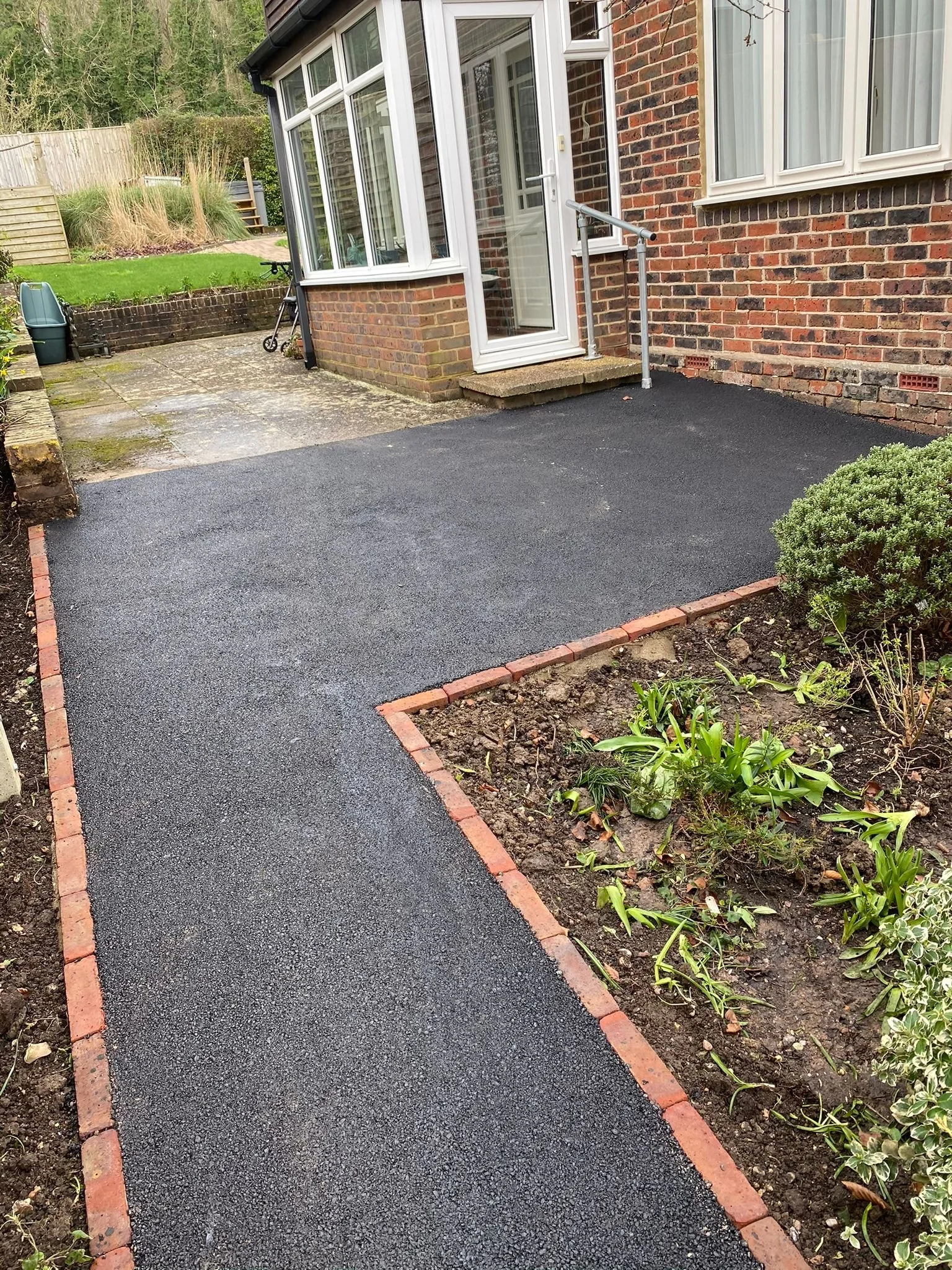 A newly paved black asphalt walkway leading to a house with a brick exterior and a white door with glass panes. To the right, there's a garden bed with green plants, bordered by red bricks. In the background, there's a patio area with concrete slabs,