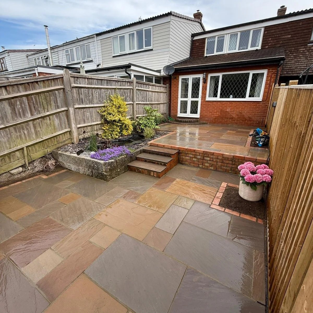 A backyard with a stone patio, steps leading to a raised deck, a small garden bed with plants, and pink flowers in a white pot. Wooden fences enclose the area, and houses are visible in the background.