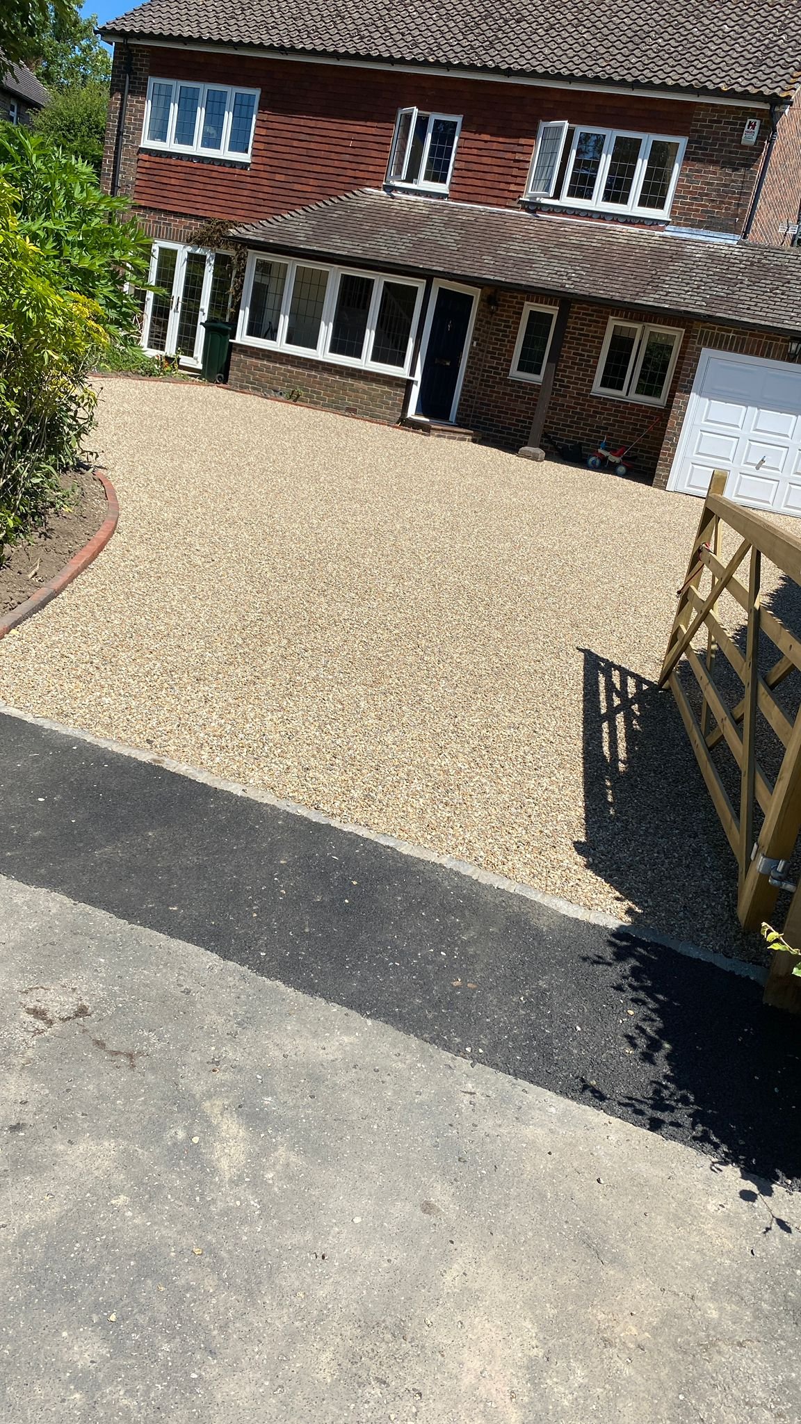 A newly laid gravel driveway in front of a two-story brick house with a brown tiled roof. The house features white-framed windows and a garage door. There is a small garden bed with green plants on the left and a wooden gate on the right, casting a s