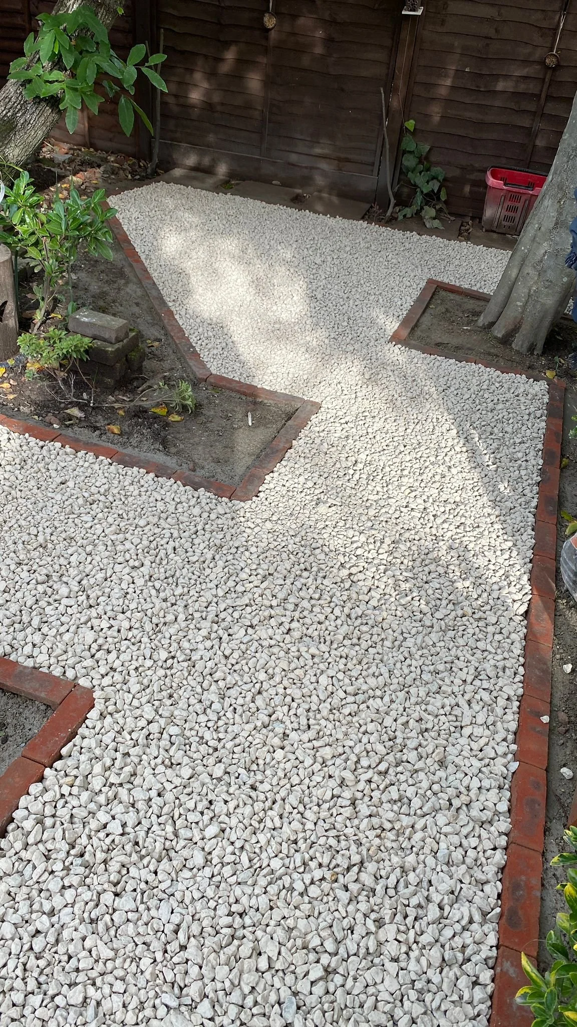 A backyard garden path made of white gravel, bordered with red bricks, with plants and small trees on the sides and a wooden fence in the background.
