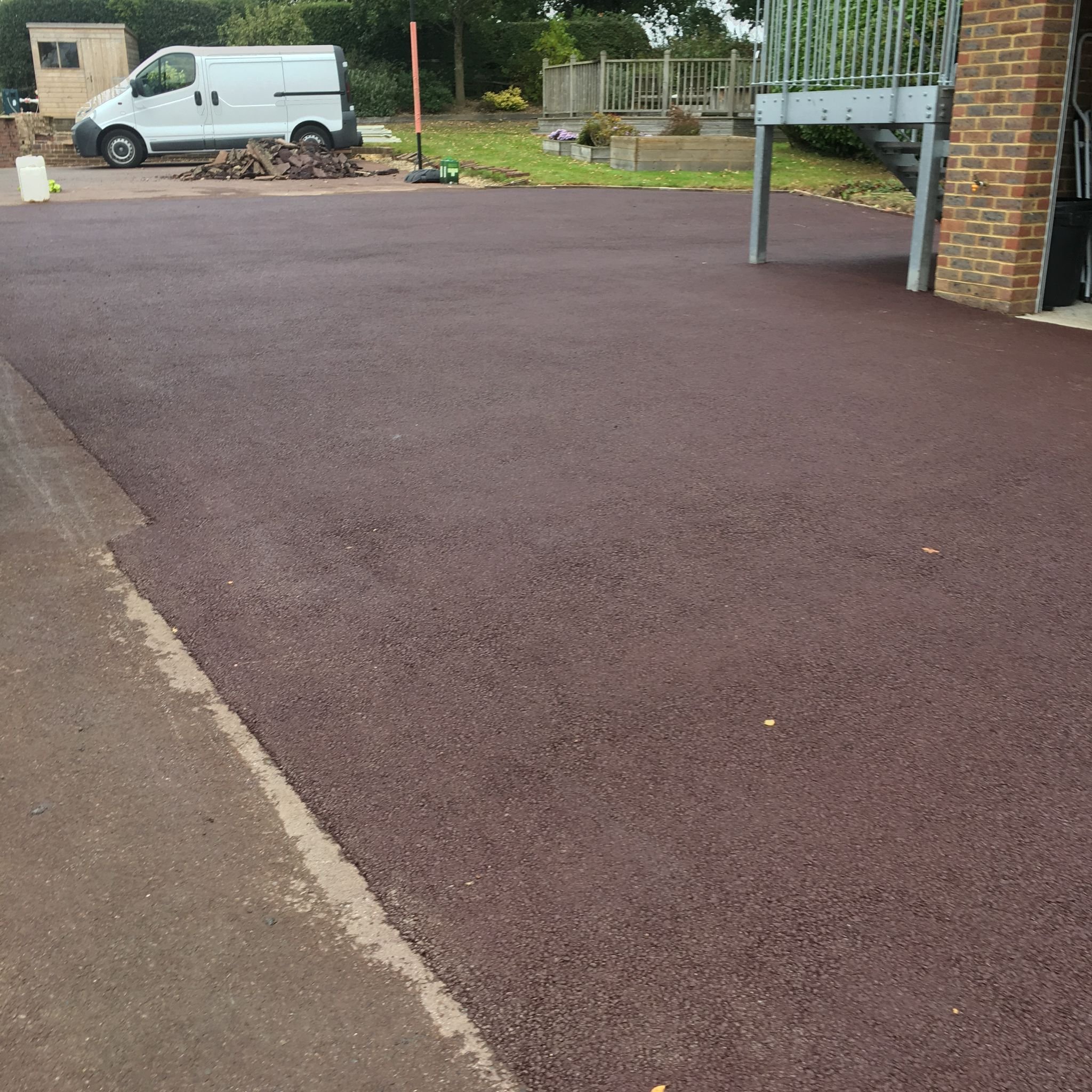Freshly paved driveway with a smooth, reddish-brown surface, adjacent to a concrete curb and a gravel pathway. In the background, there's a white van parked near a pile of rocks, alongside a grassy yard with trees and a wooden shed. A brick house wit