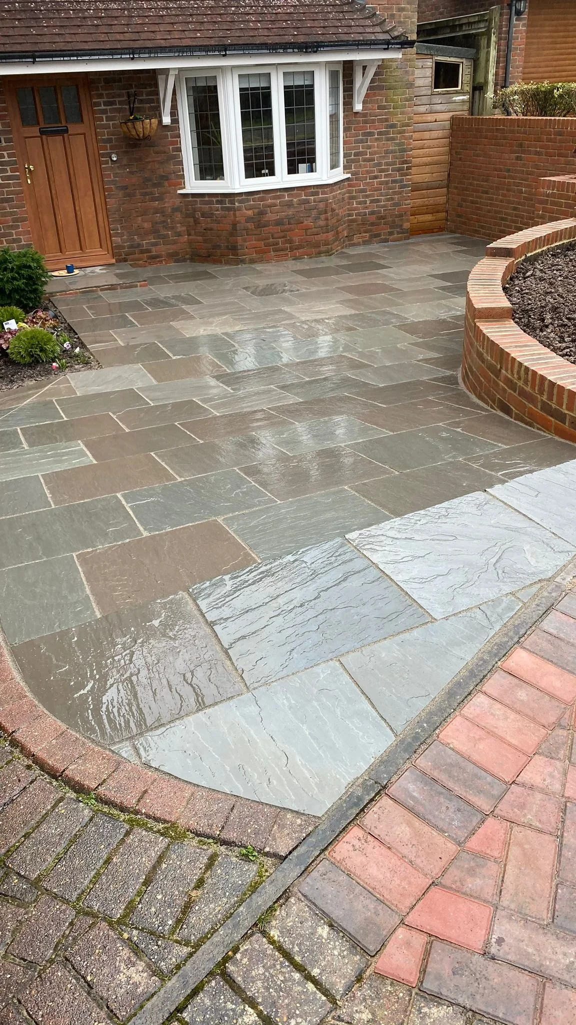 Wet stone patio in front of a brick house with bay window and flower bed to the left.